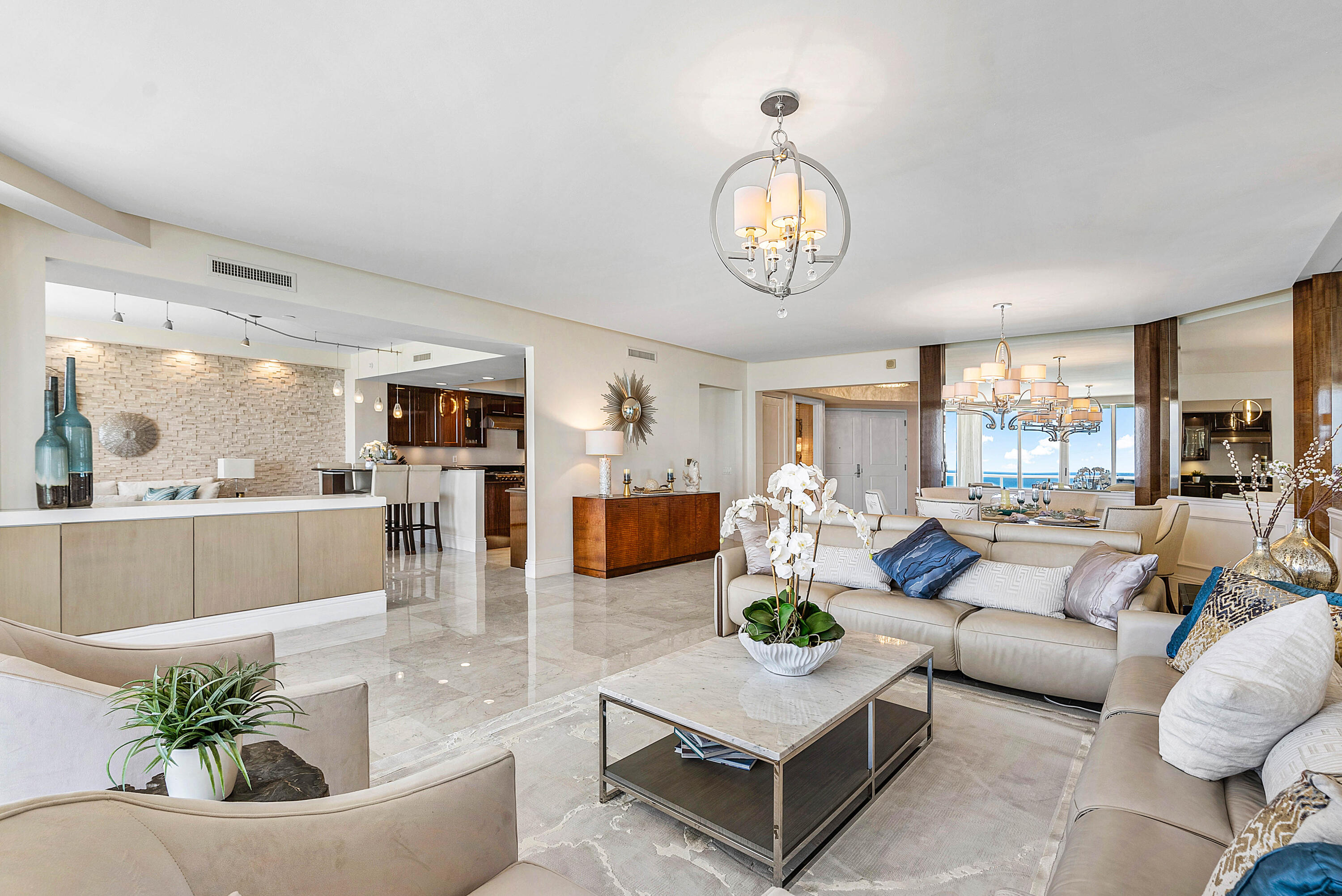 Open-plan living room with cream sofas, marble floors, and ocean views through the dining area windows.