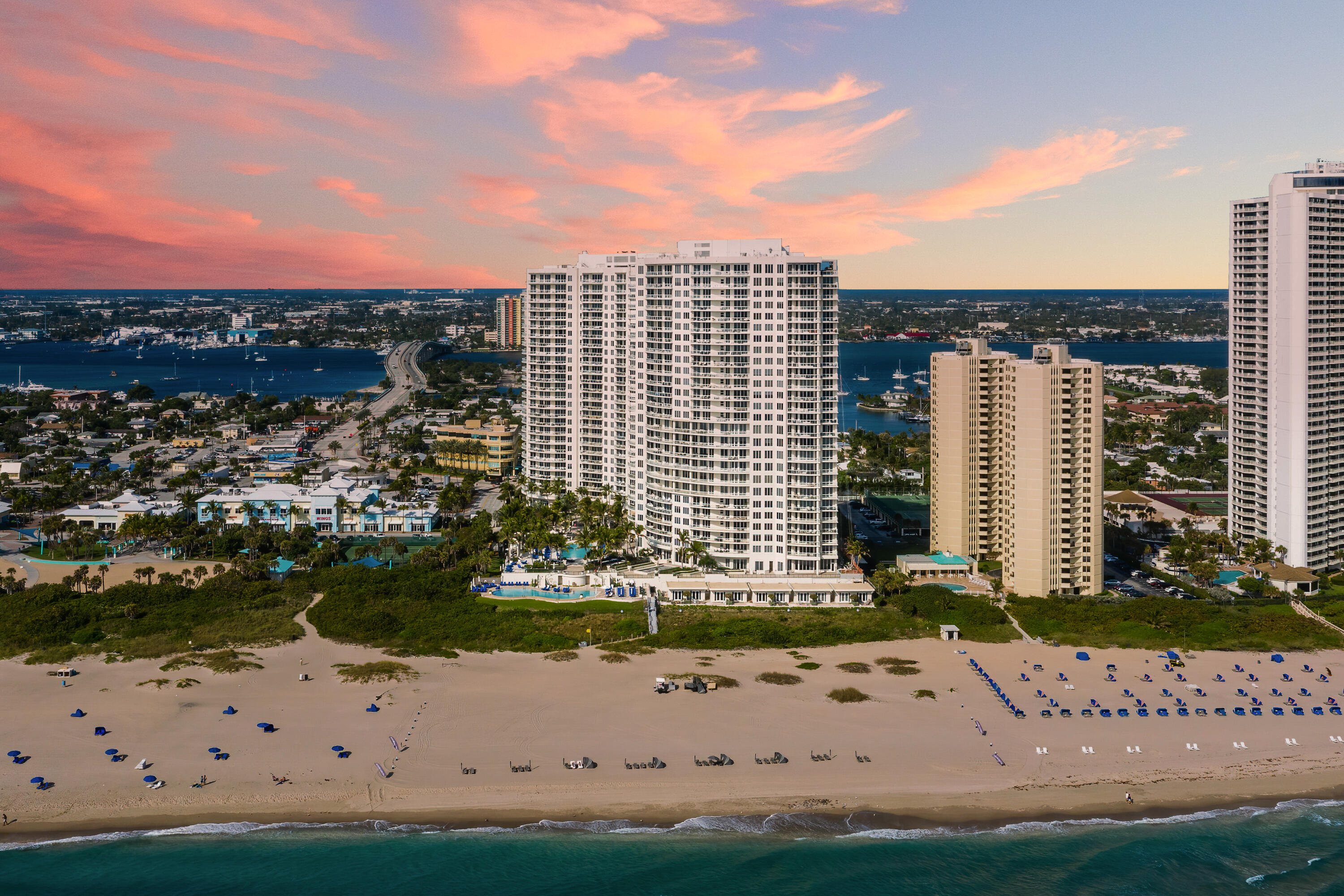 Beachfront condominium tower beside a sandy beach, ocean, inlet, and bridge at sunset.