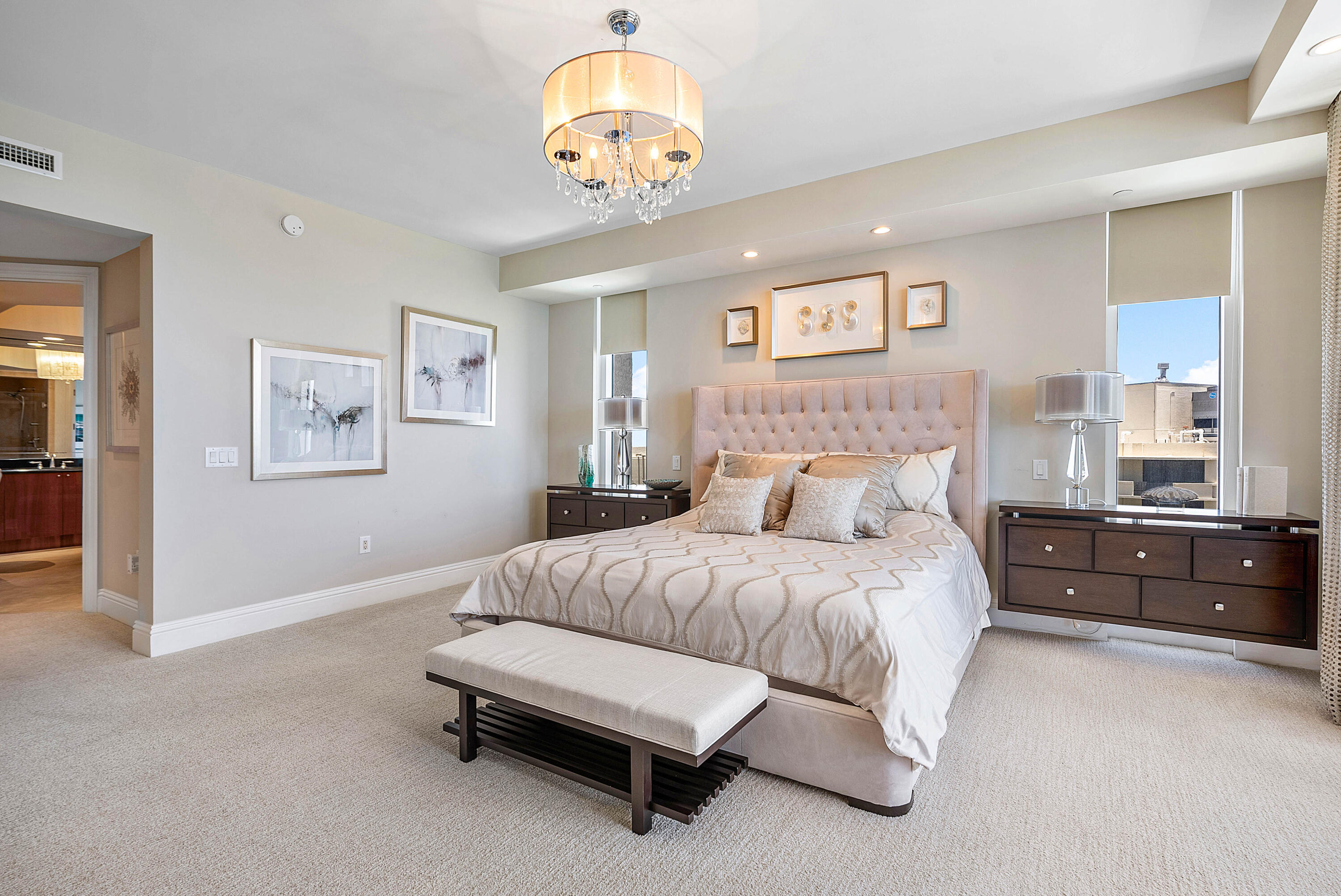 Staged bedroom with a tufted headboard, cream bedding, chandelier, and windows overlooking rooftops.