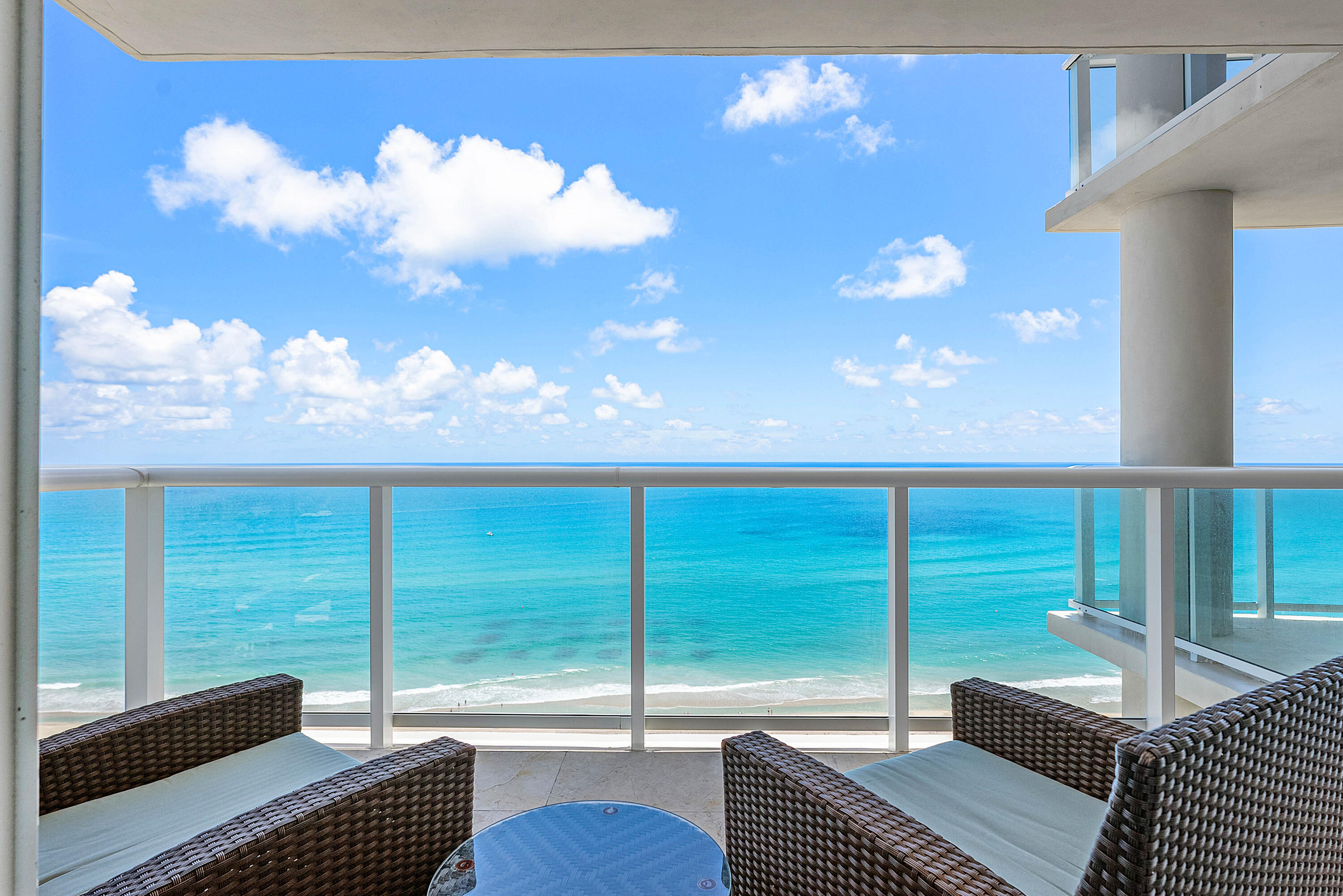 Furnished balcony overlooking turquoise ocean and beach, with wicker seating and a glass railing.