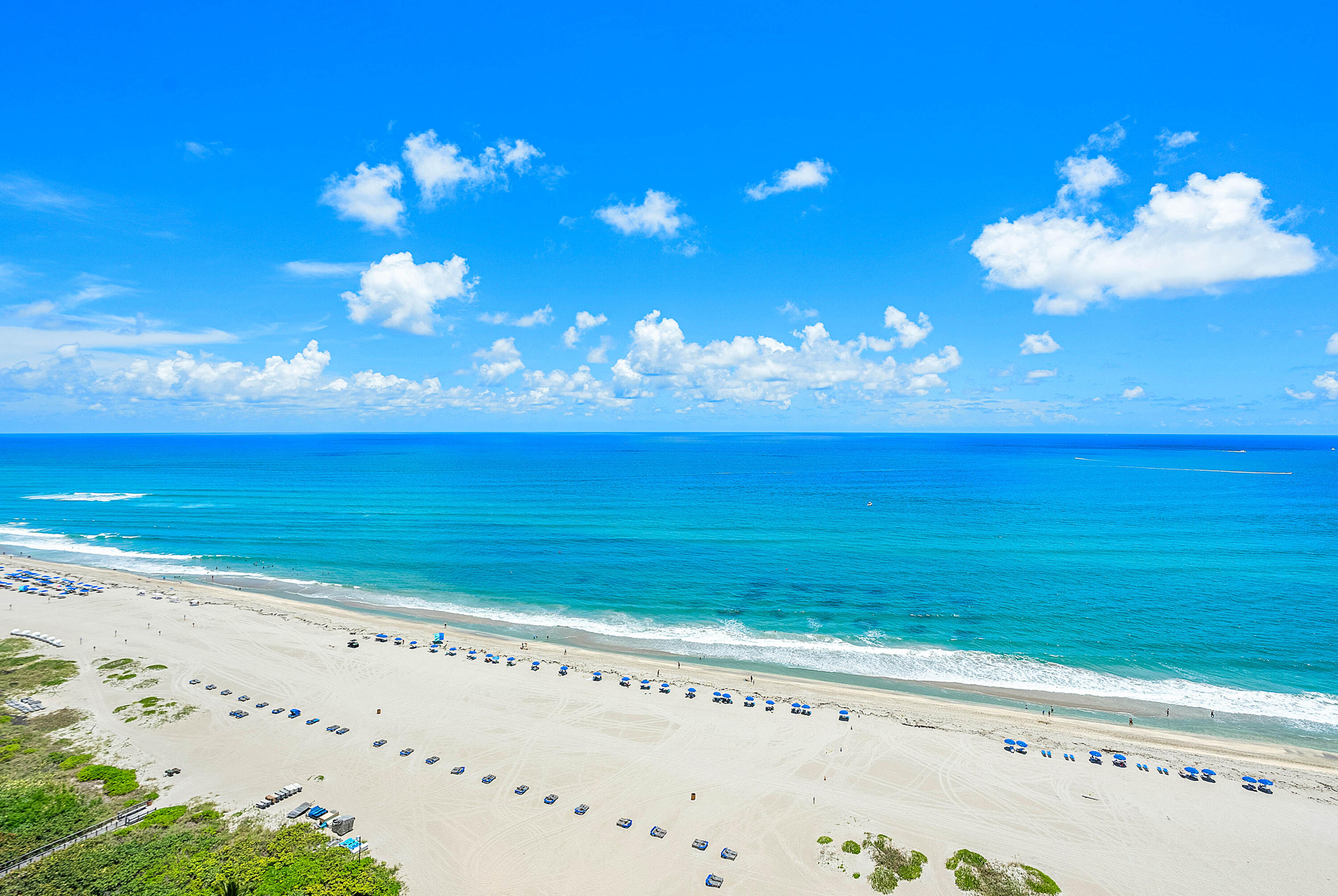 Turquoise ocean and sandy beach lined with blue umbrellas beneath a cloud-filled sky.