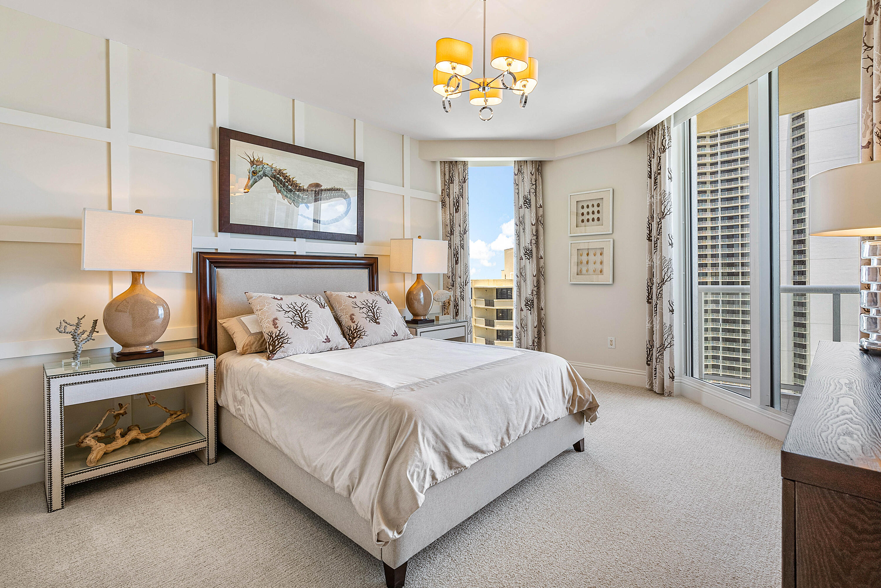 Bedroom with a white bed, seahorse artwork, and floor-to-ceiling windows overlooking neighboring towers.