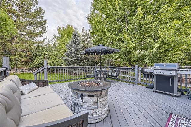 Wooden deck with cushioned seating, a stone fire pit, patio umbrella, and grill among mature trees.