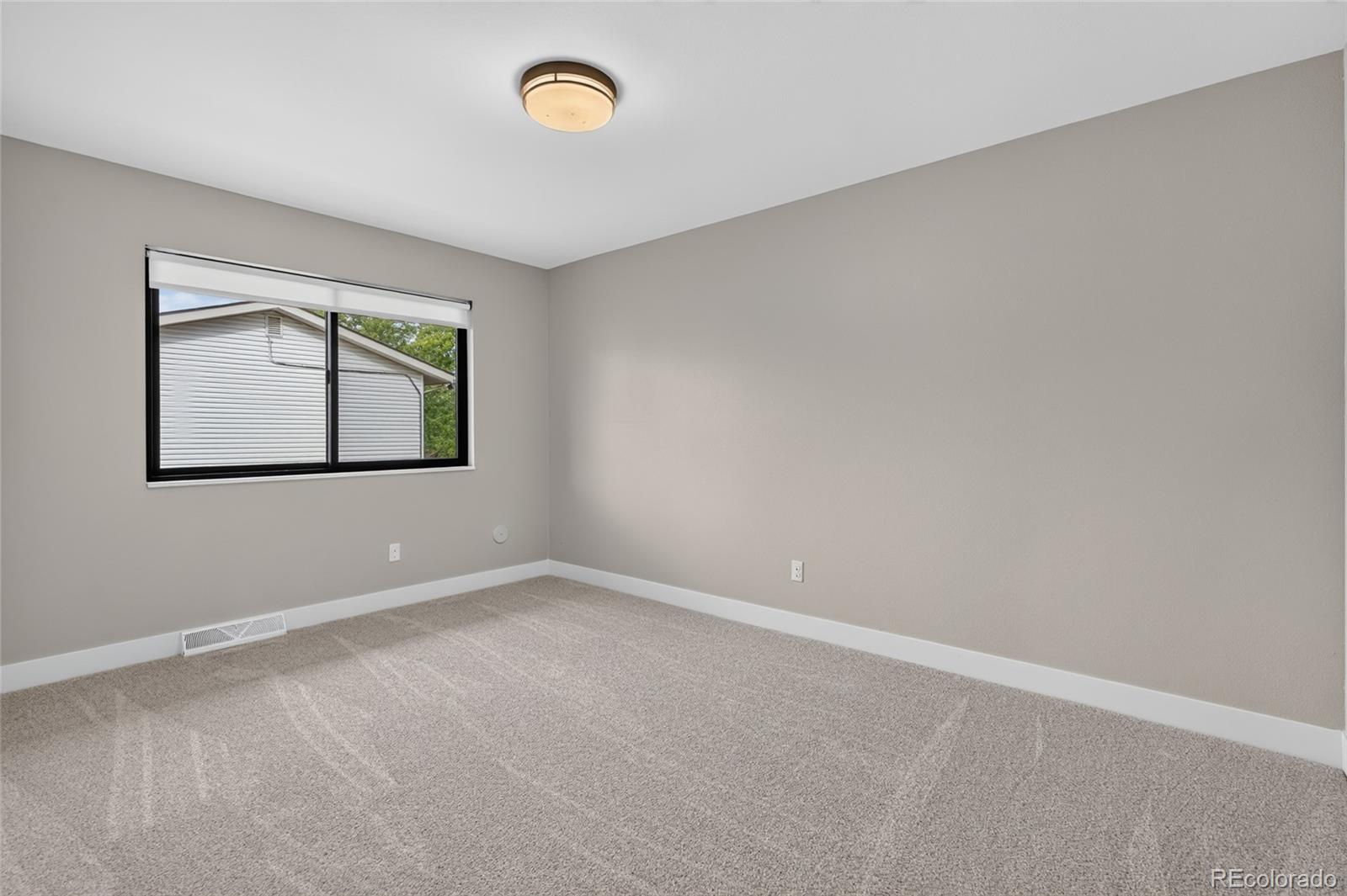 Vacant bedroom with beige carpet, gray walls, and a window overlooking a neighboring house.
