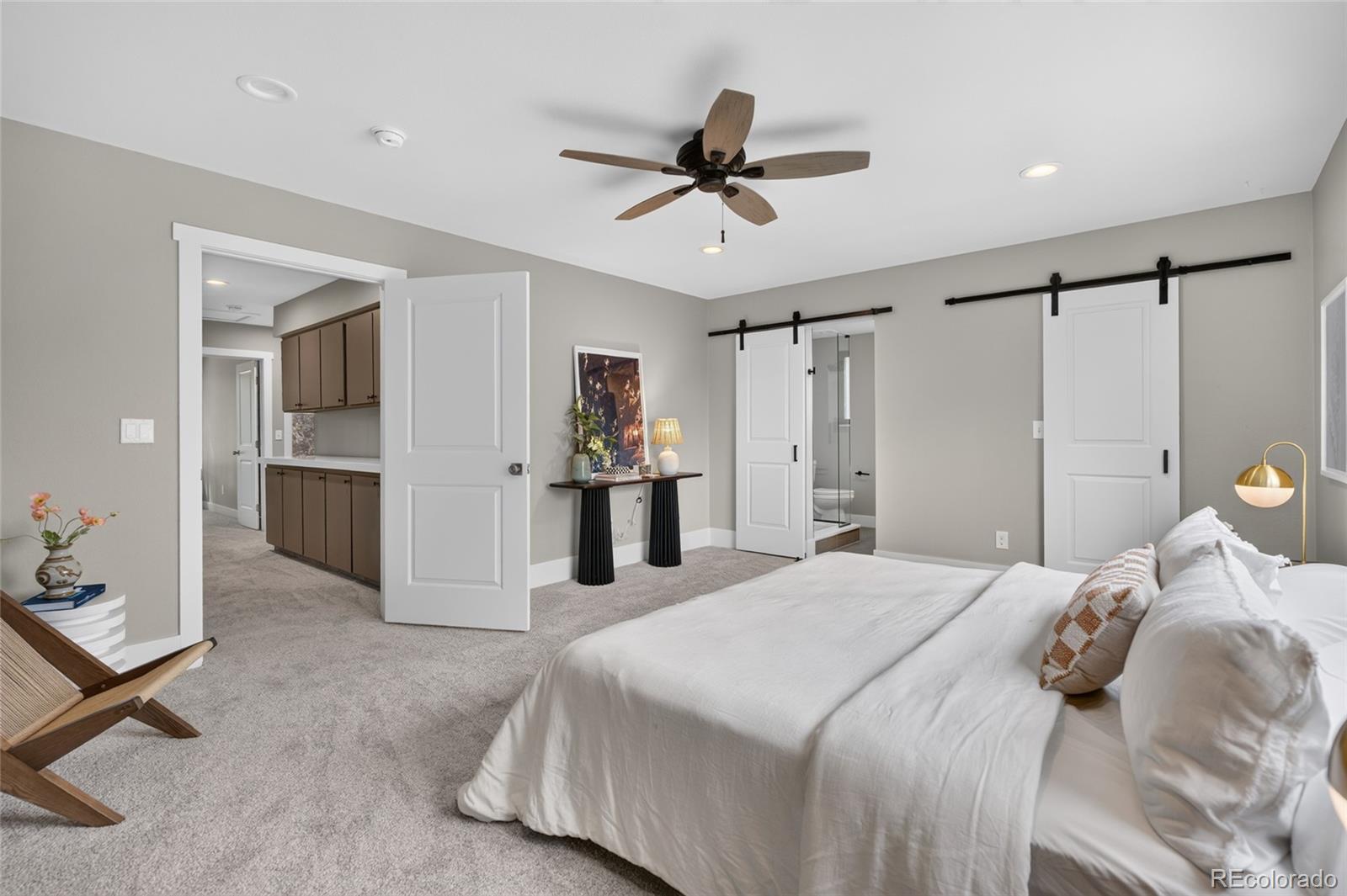 Staged bedroom with a white bed, ceiling fan, and sliding barn doors beside an open bathroom doorway.