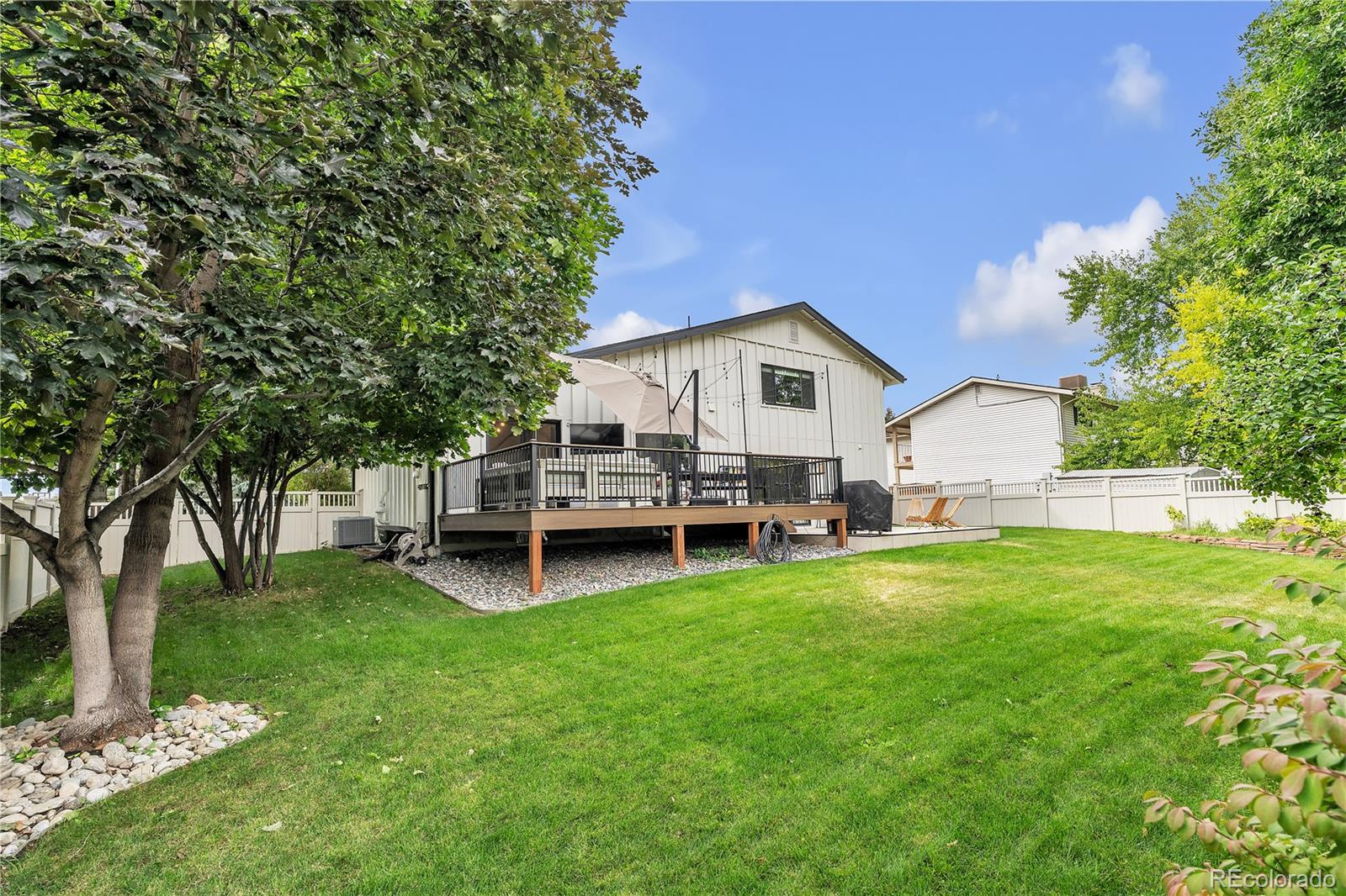 Home’s white-sided rear exterior and raised deck overlook a spacious lawn shaded by mature trees.