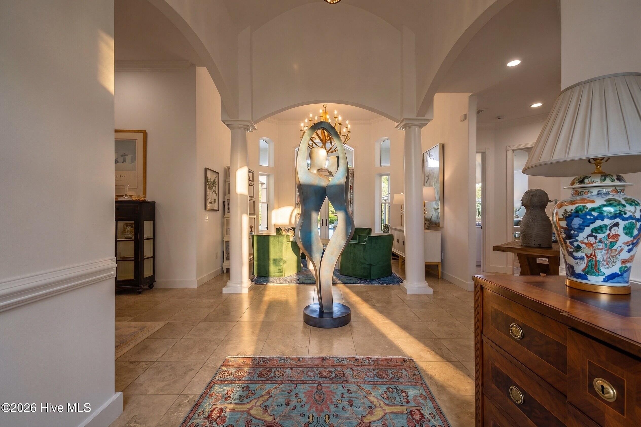 Foyer with a tall abstract sculpture, arched columns, green armchairs, and sunlight across the tiled floor.