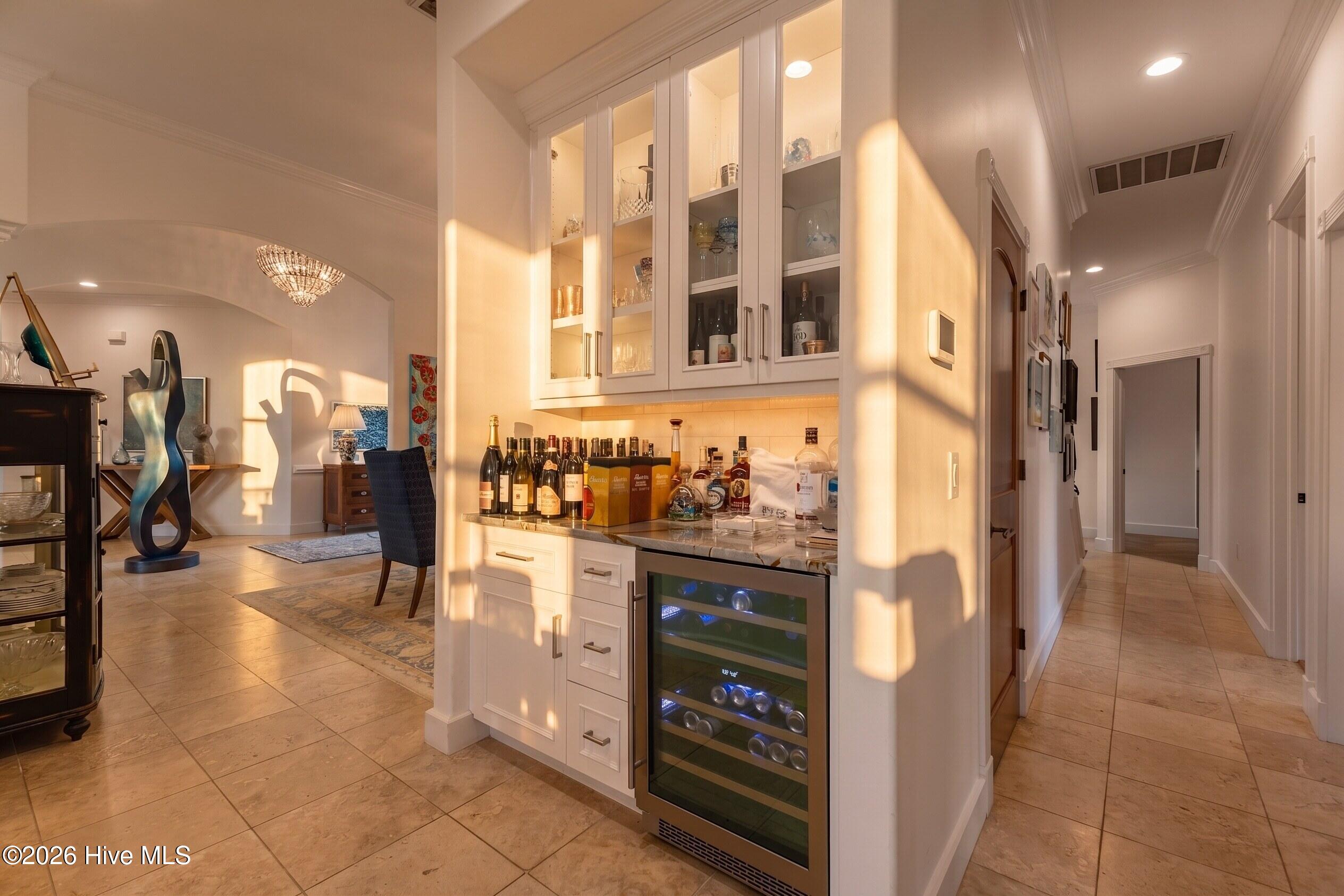 Built-in white bar with glass-front cabinets and wine refrigerator beside a sunlit sitting area.