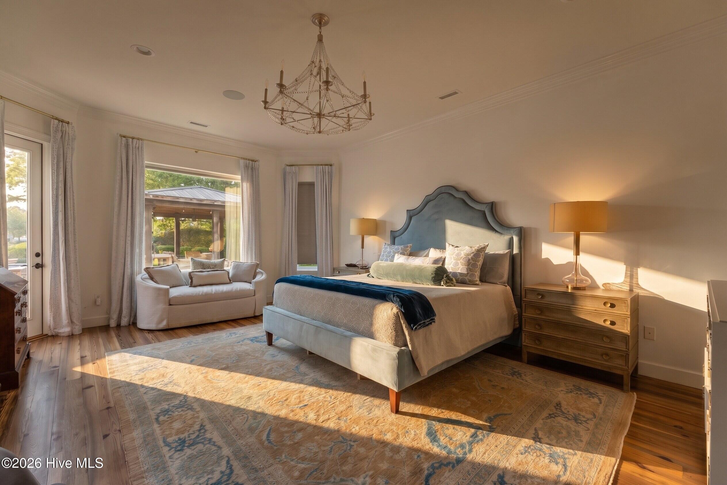 Furnished bedroom with an upholstered bed, chandelier, and window-side sitting area in warm sunlight.