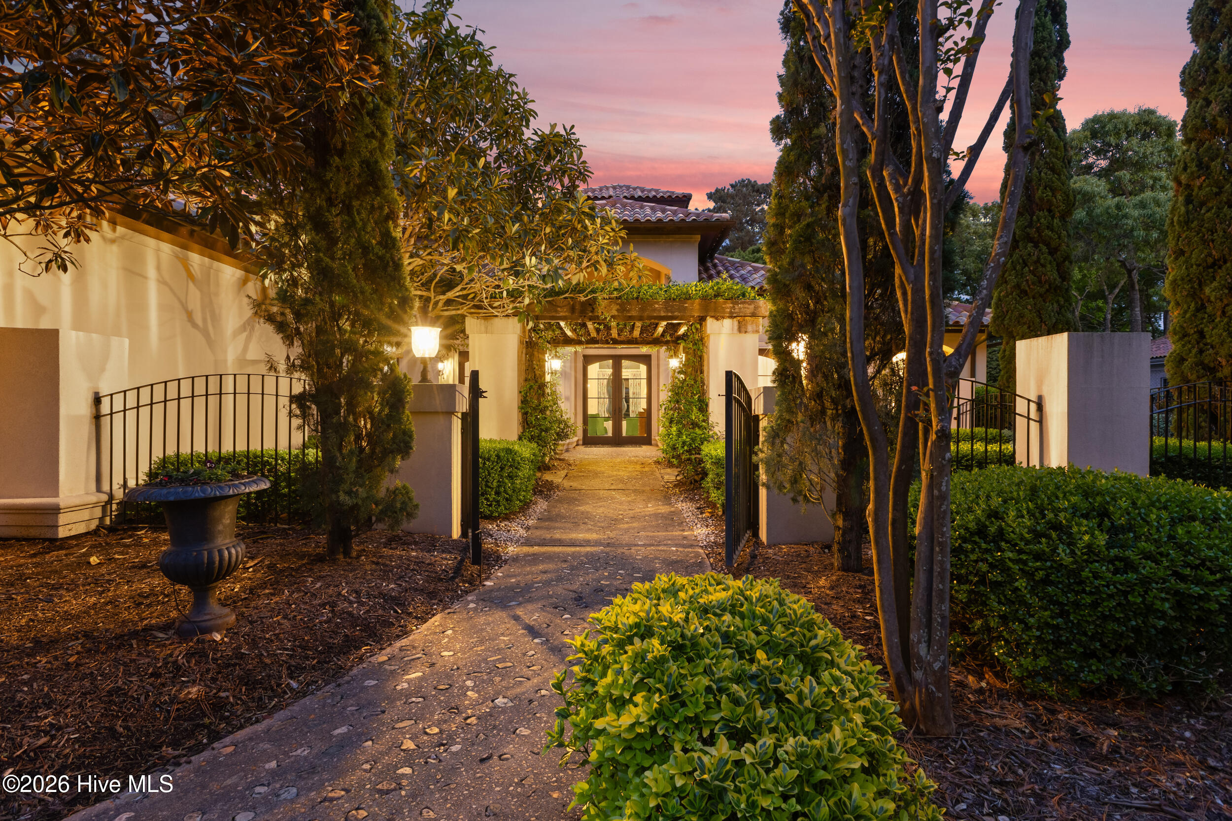 Home entrance with an open wrought-iron gate, stone path, and pergola at dusk.