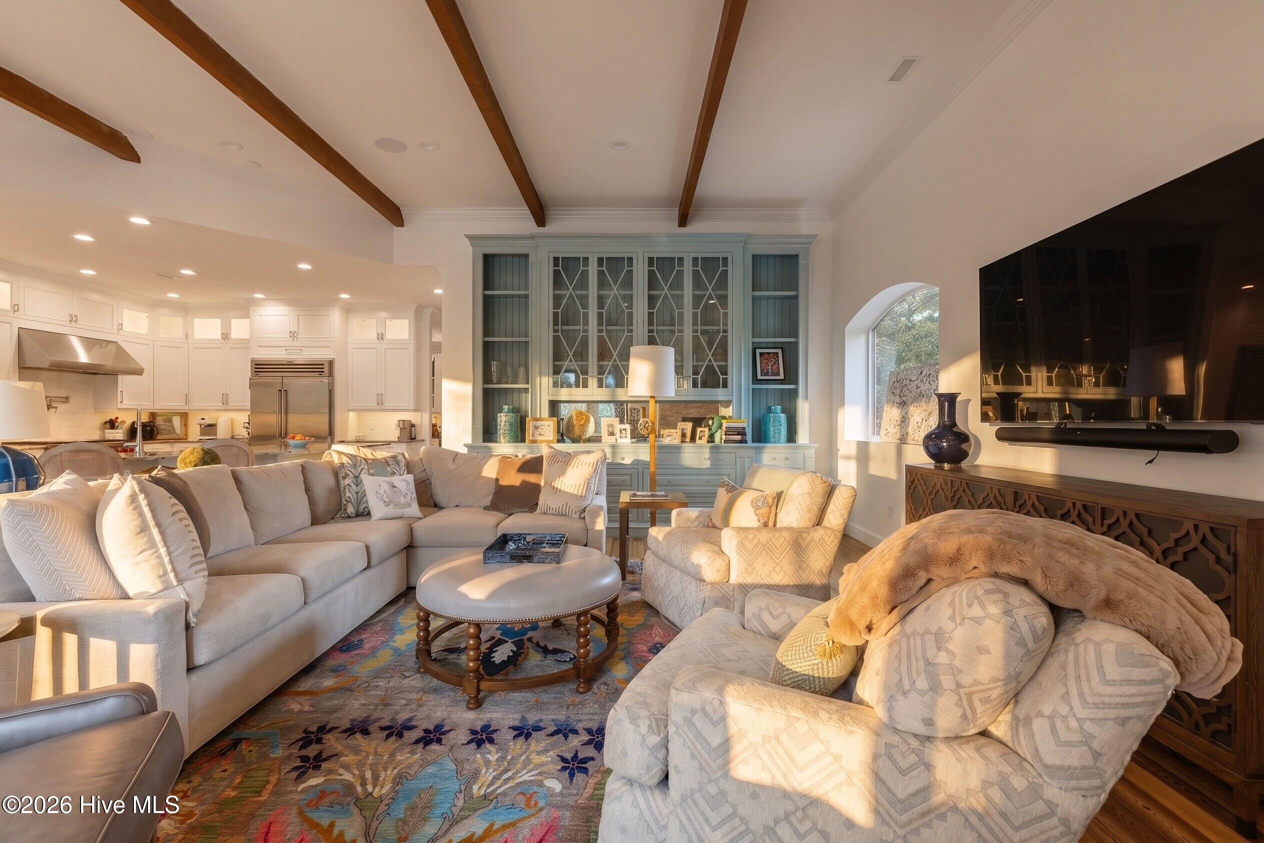 Living room with wood ceiling beams, blue built-in cabinetry, pale seating, and a colorful patterned rug.