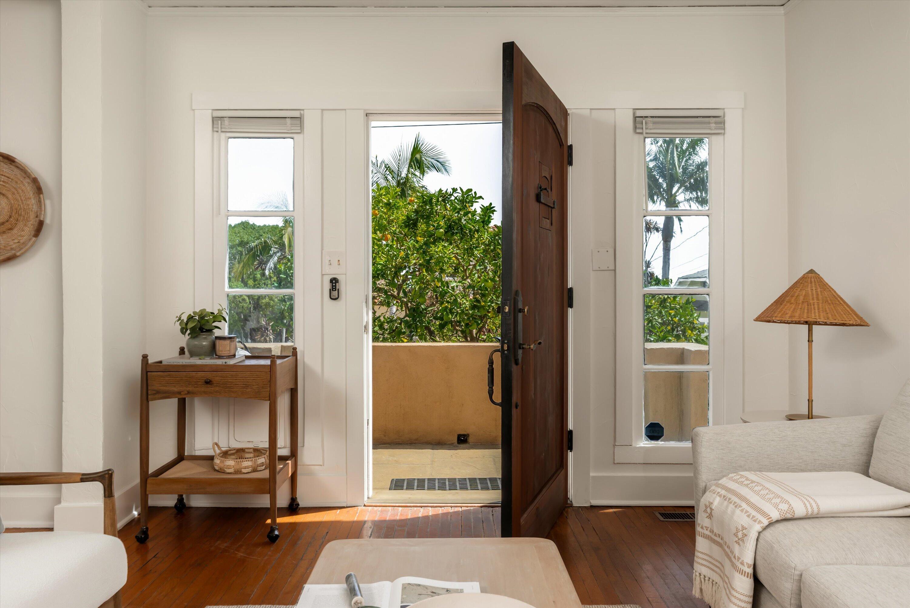 Living room with an open dark wood front door, flanking windows, and leafy greenery outside.