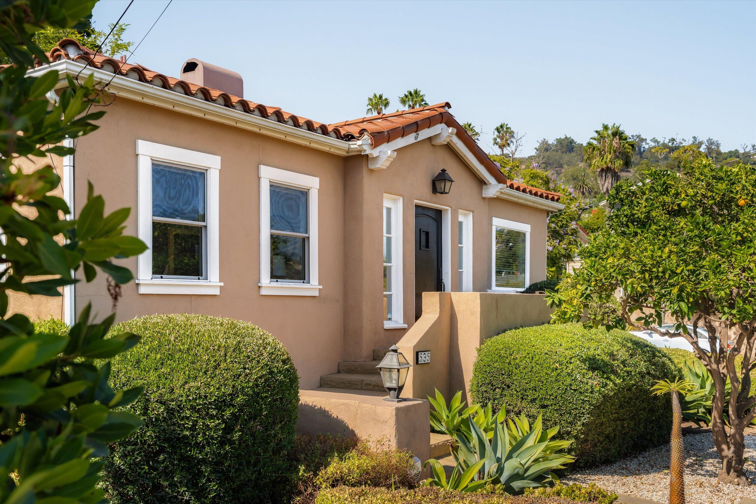 Tan stucco home with terracotta tile roof, recessed entry, and mature landscaped front yard.