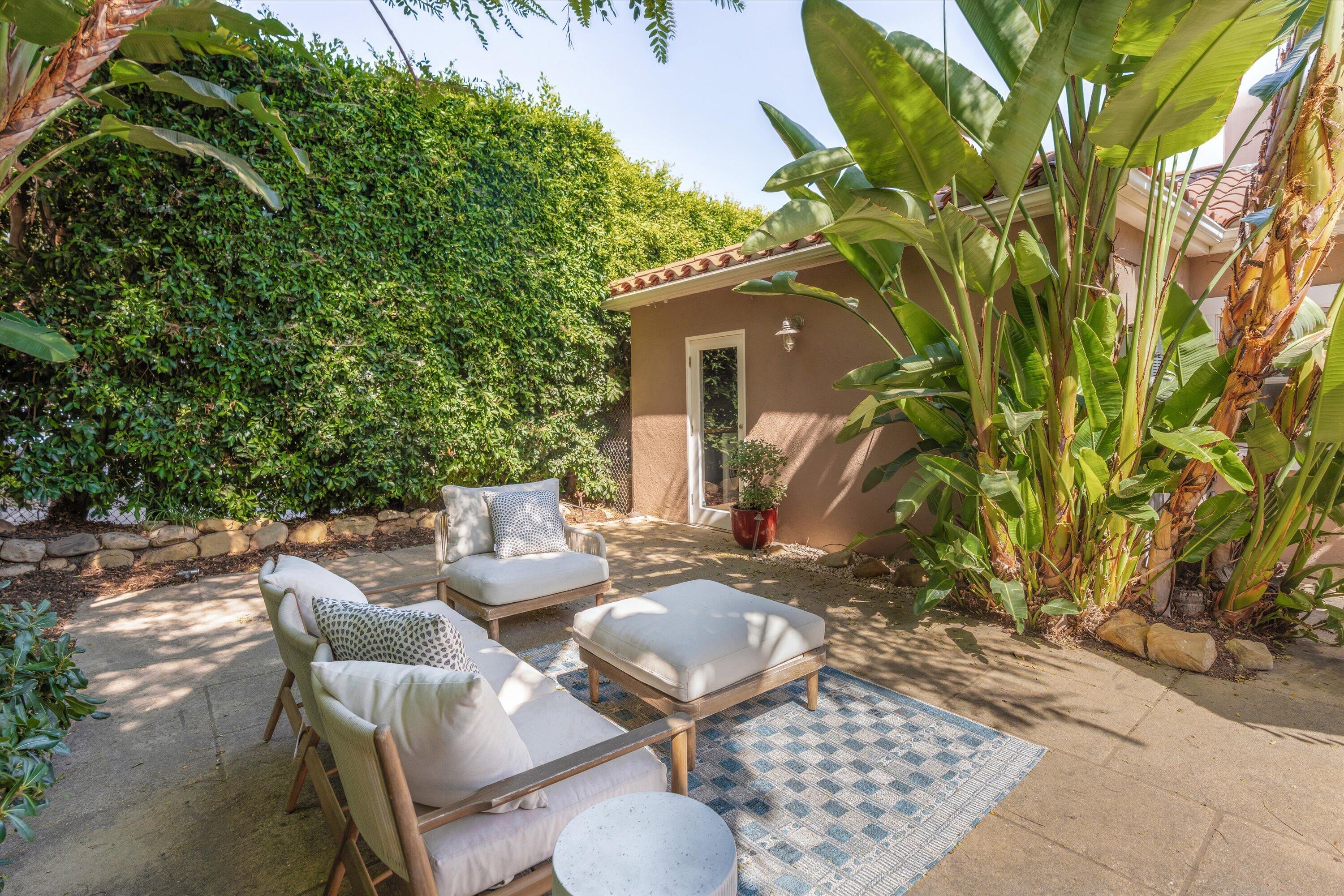 Furnished patio with white-cushioned lounge chairs, a patterned rug, and tall tropical plants.
