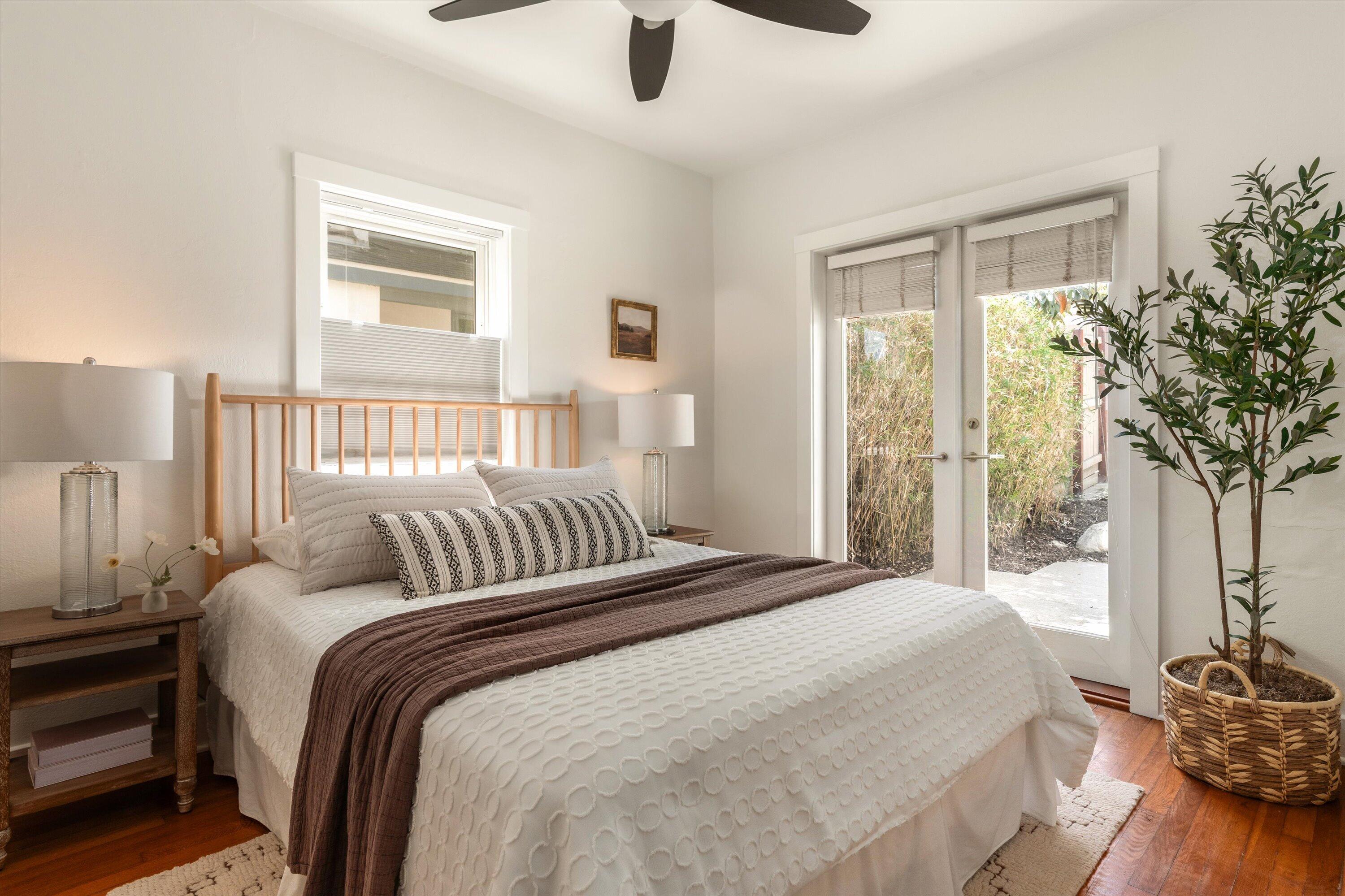 Bright bedroom with a white bed, dark throw, French doors to a patio, and hardwood floors.
