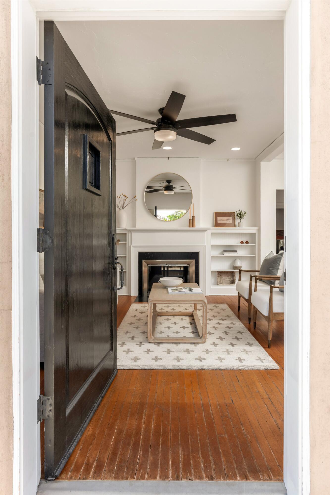 Staged living room with a white fireplace, built-in shelves, warm wood floors, and an open dark front door.