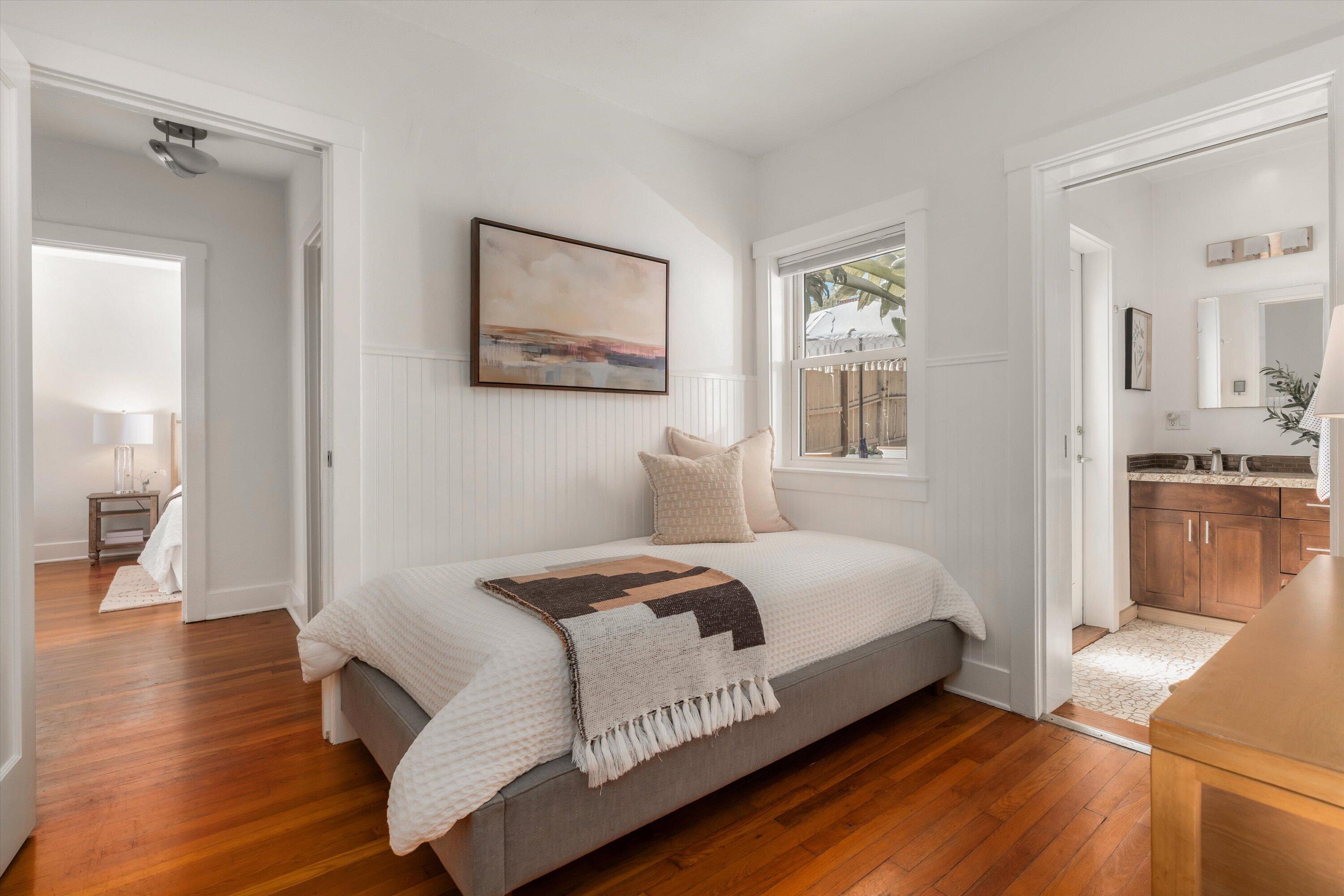 Staged bedroom with a white bed, hardwood floors, and doorways to a bathroom and adjoining bedroom.