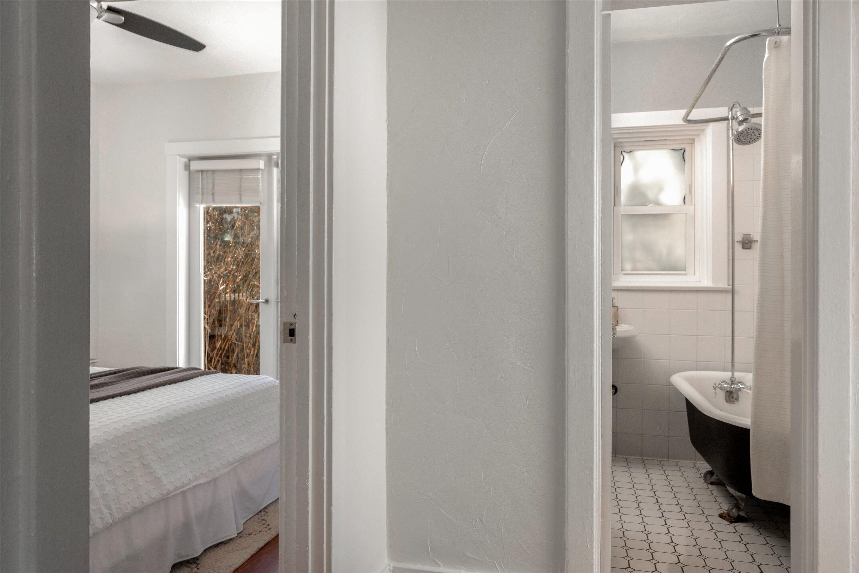Bedroom and adjoining bathroom with a black clawfoot tub and white hexagonal tile floor.