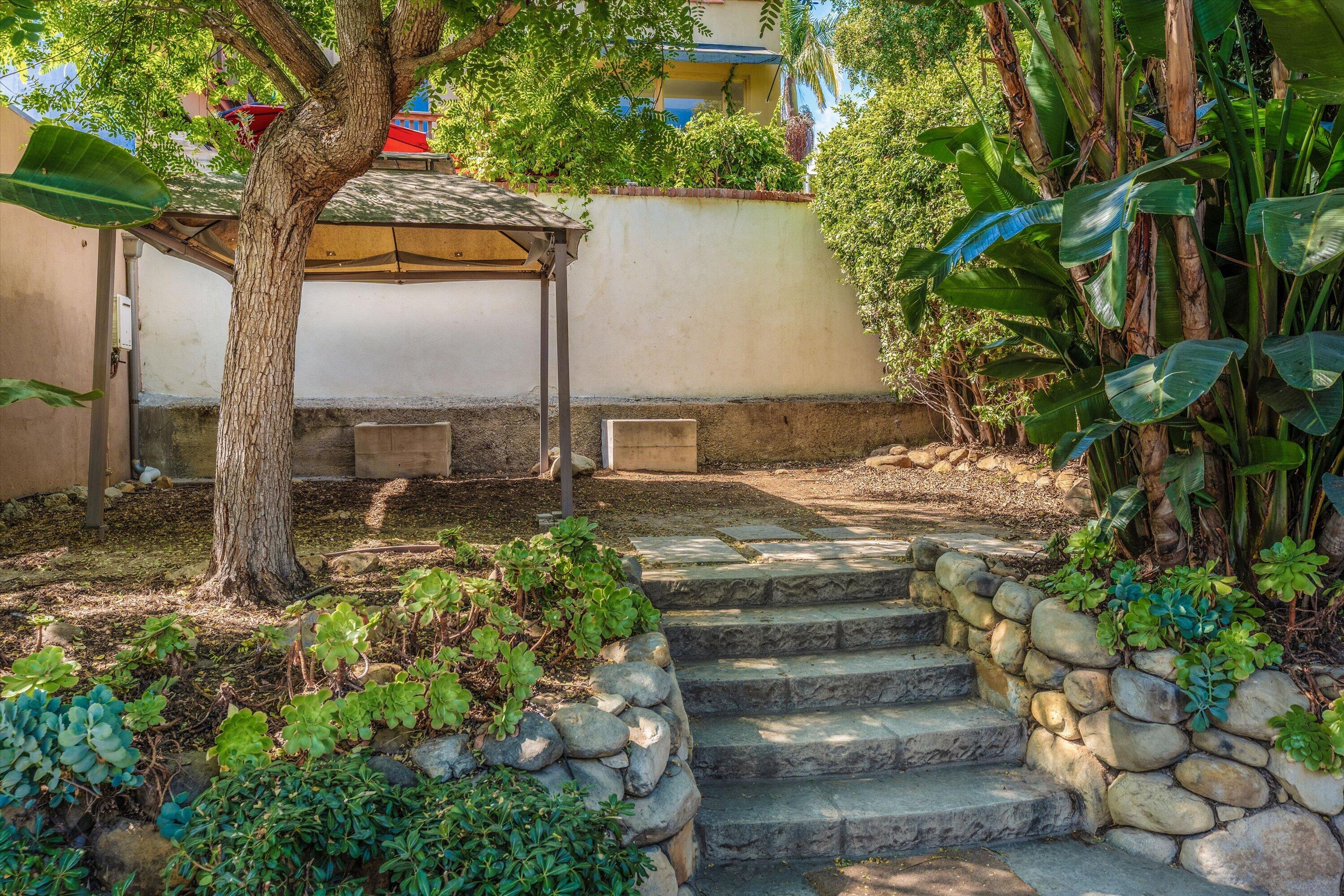 Landscaped backyard with stone steps, rounded stone walls, and a pergola shaded by mature trees.