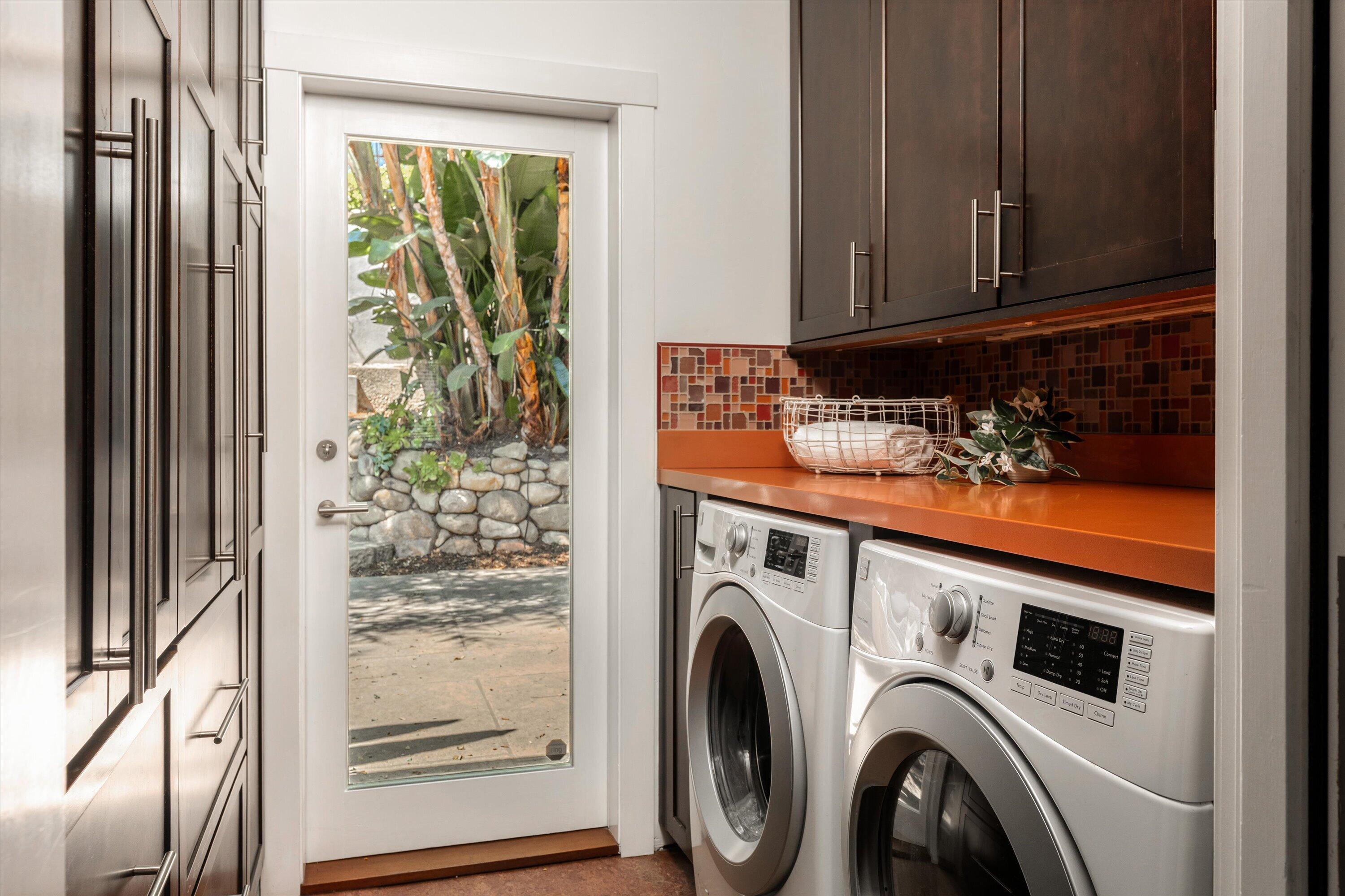 Laundry room with front-loading washer and dryer, orange counter, and glass door to a leafy garden.