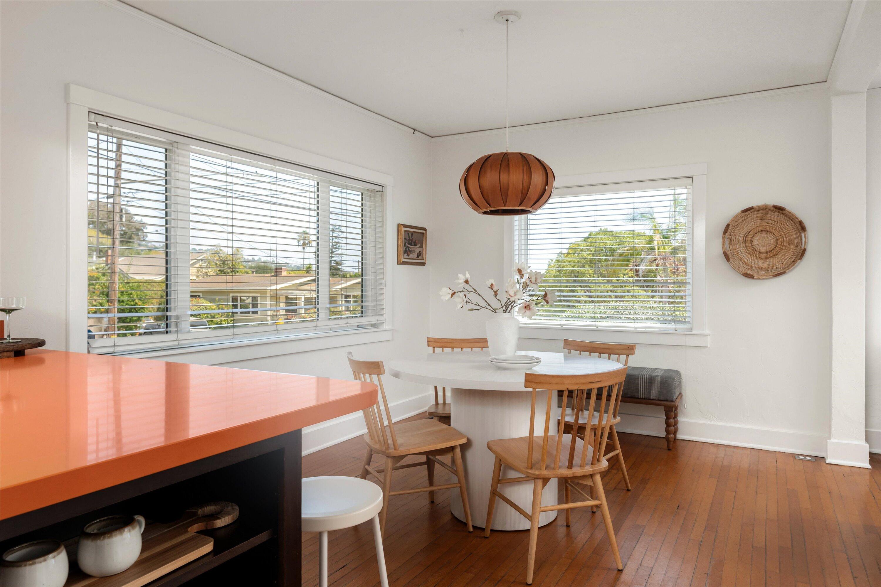 Dining area with a round white table, wooden chairs, and large windows beside an orange kitchen island.