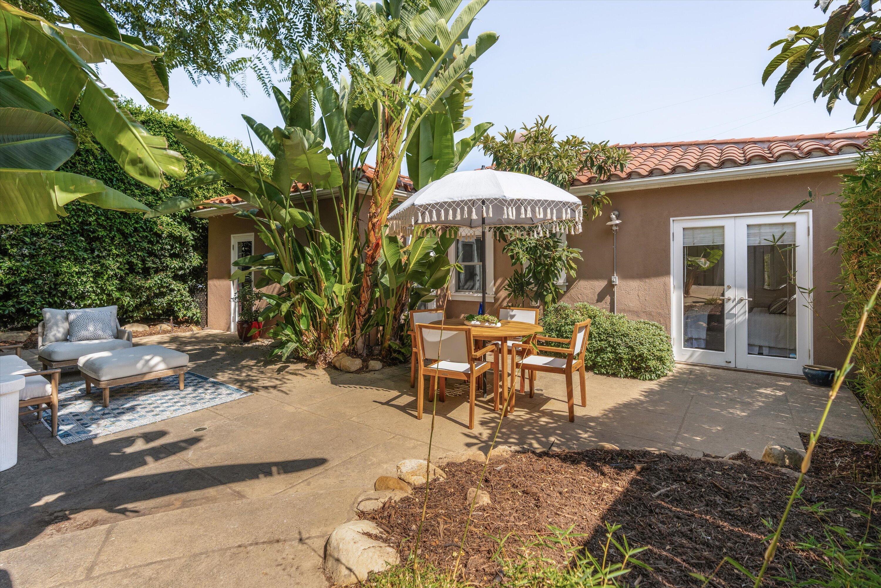 Backyard patio with outdoor dining beneath a fringed umbrella and lounge seating among tropical plants.