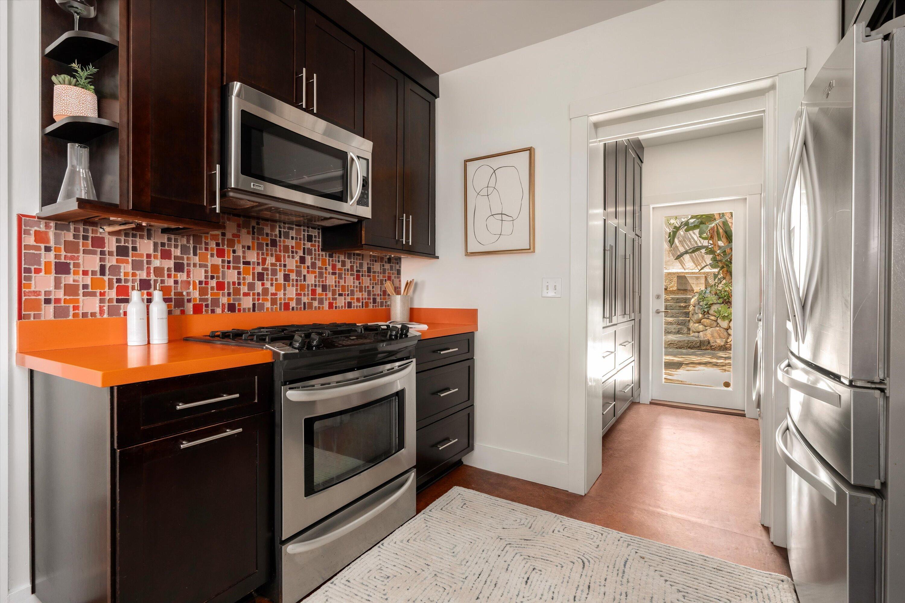 Kitchen with dark cabinets, stainless appliances, orange countertops, and a colorful mosaic backsplash.