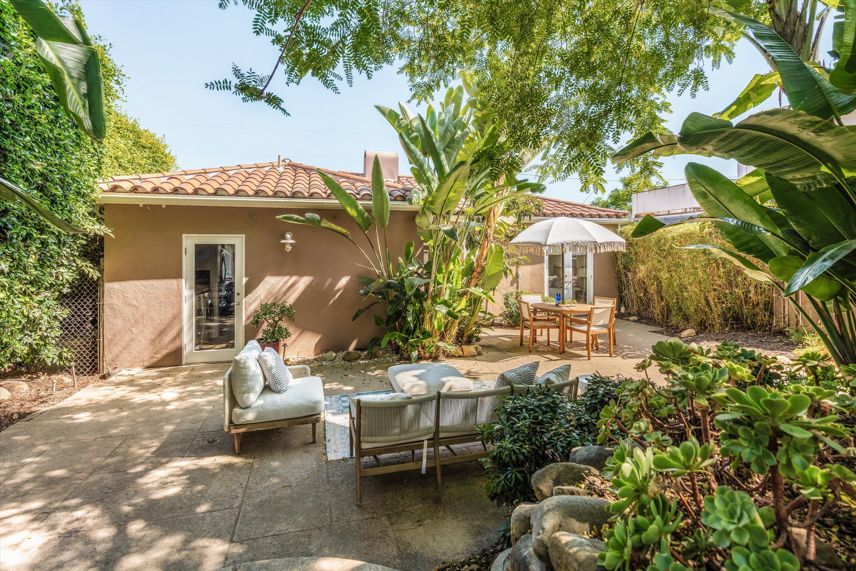 Backyard patio with lounge seating, a dining table under a fringed umbrella, and lush tropical landscaping.
