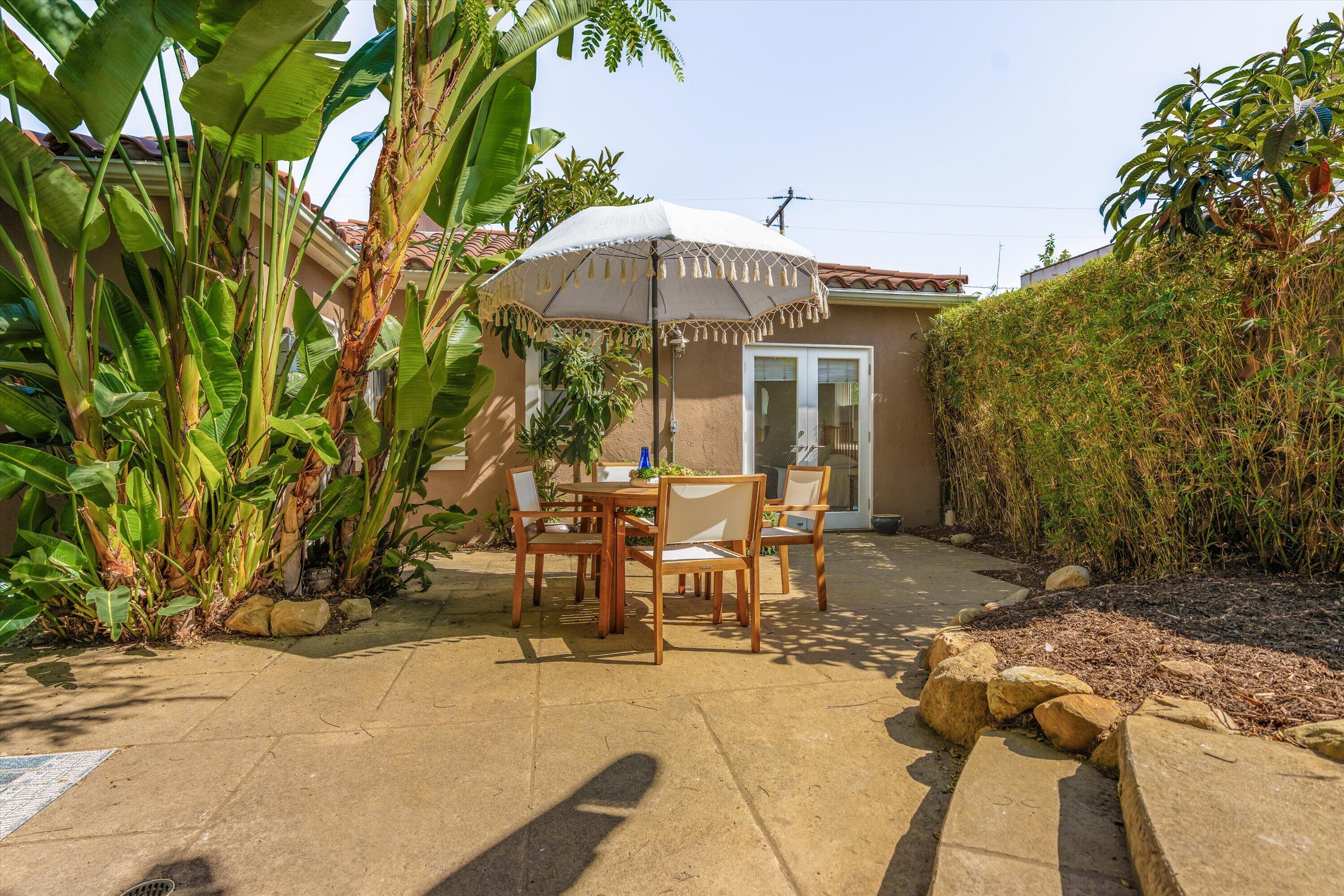 Backyard patio with a dining set beneath a fringed umbrella, surrounded by tropical plants and a tall hedge.