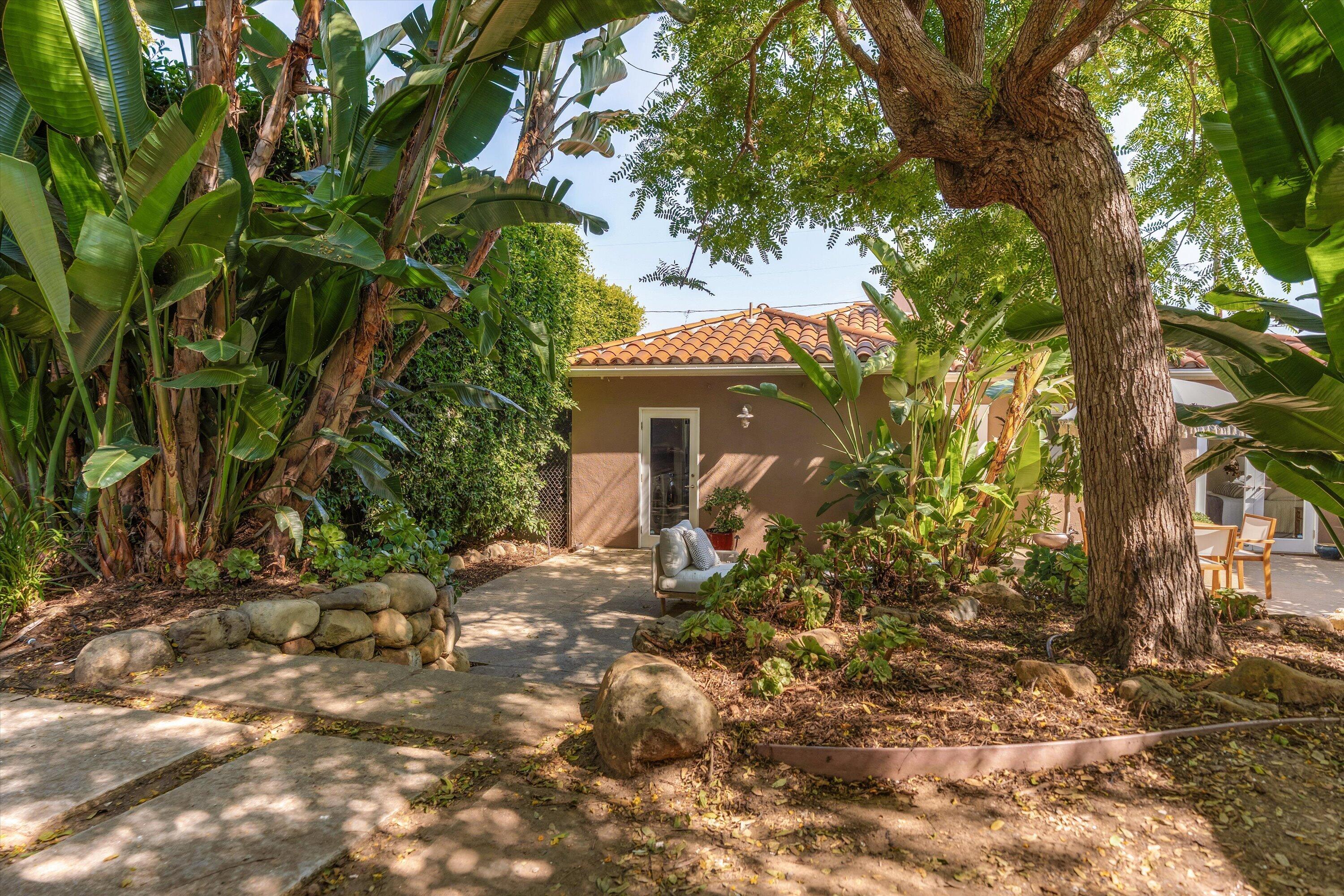 Garden path winds through lush tropical landscaping beside a terracotta-roofed home and large shade tree.