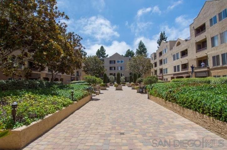 Landscaped courtyard walkway between multi-story residential buildings, bordered by raised planting beds and mature trees.