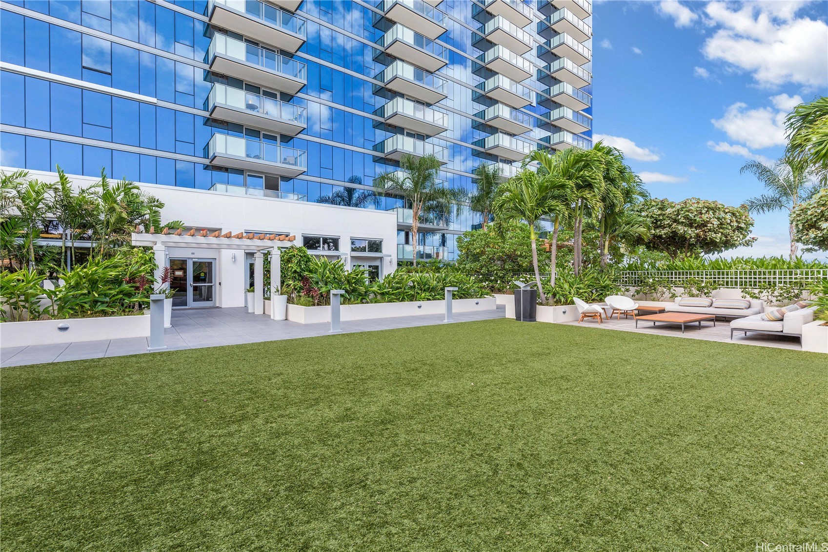 Condominium courtyard with tropical landscaping, a turf lawn, and outdoor lounge seating beside a glass high-rise.
