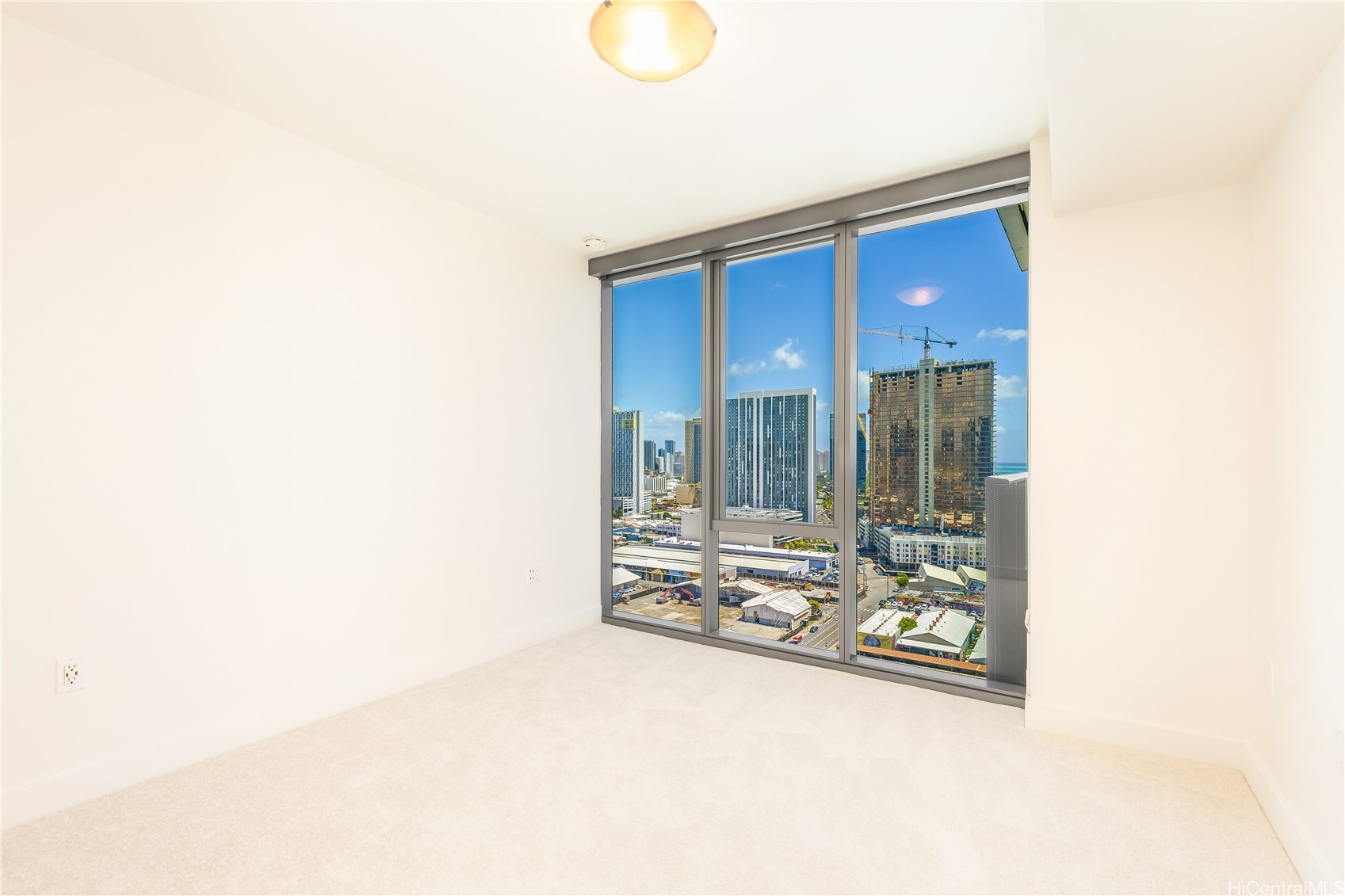 Empty carpeted room with floor-to-ceiling windows overlooking Honolulu high-rises and a glimpse of the ocean.