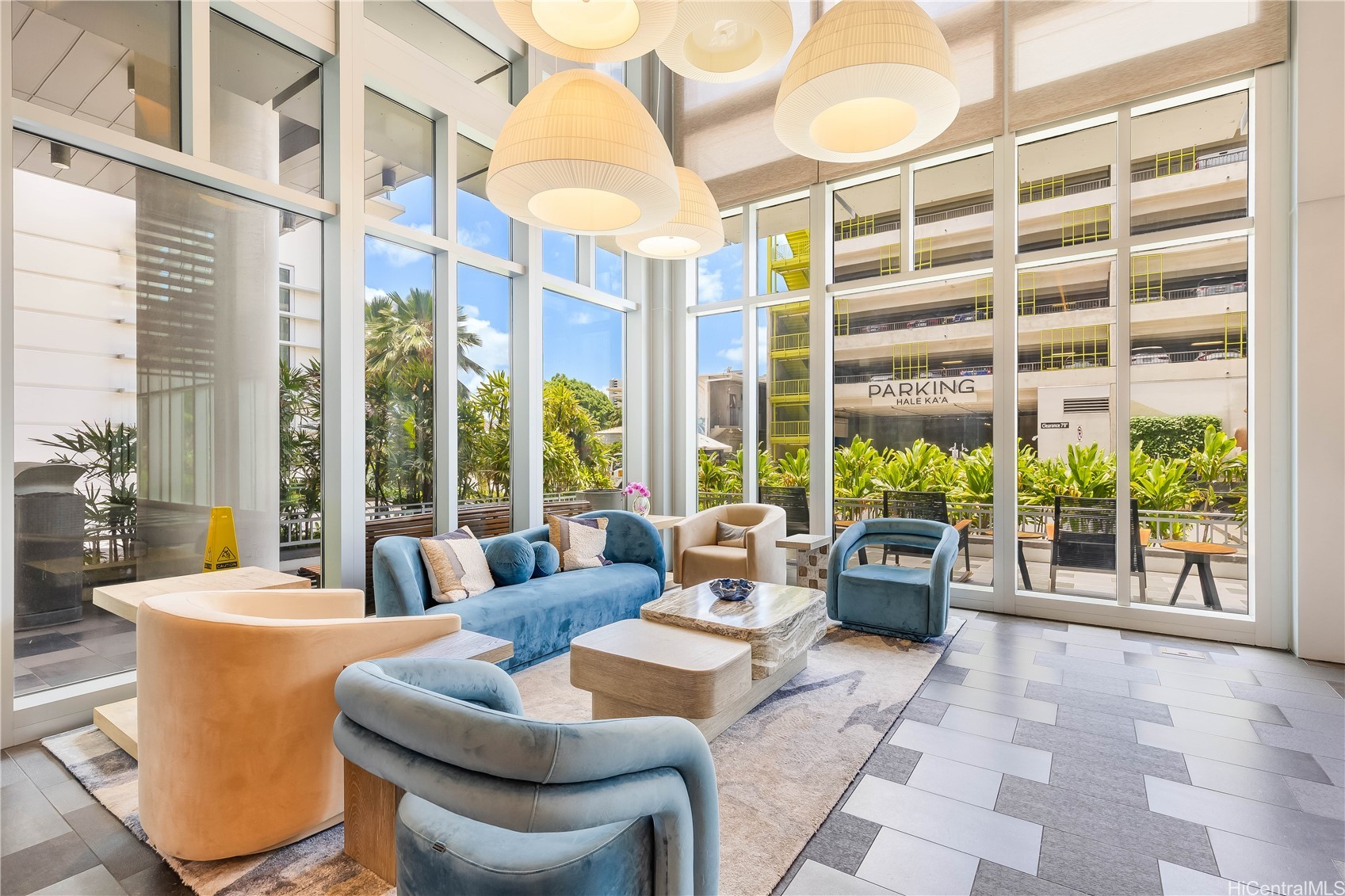 Furnished lounge with blue and peach seating, oversized pendant lights, and floor-to-ceiling windows overlooking tropical landscaping.