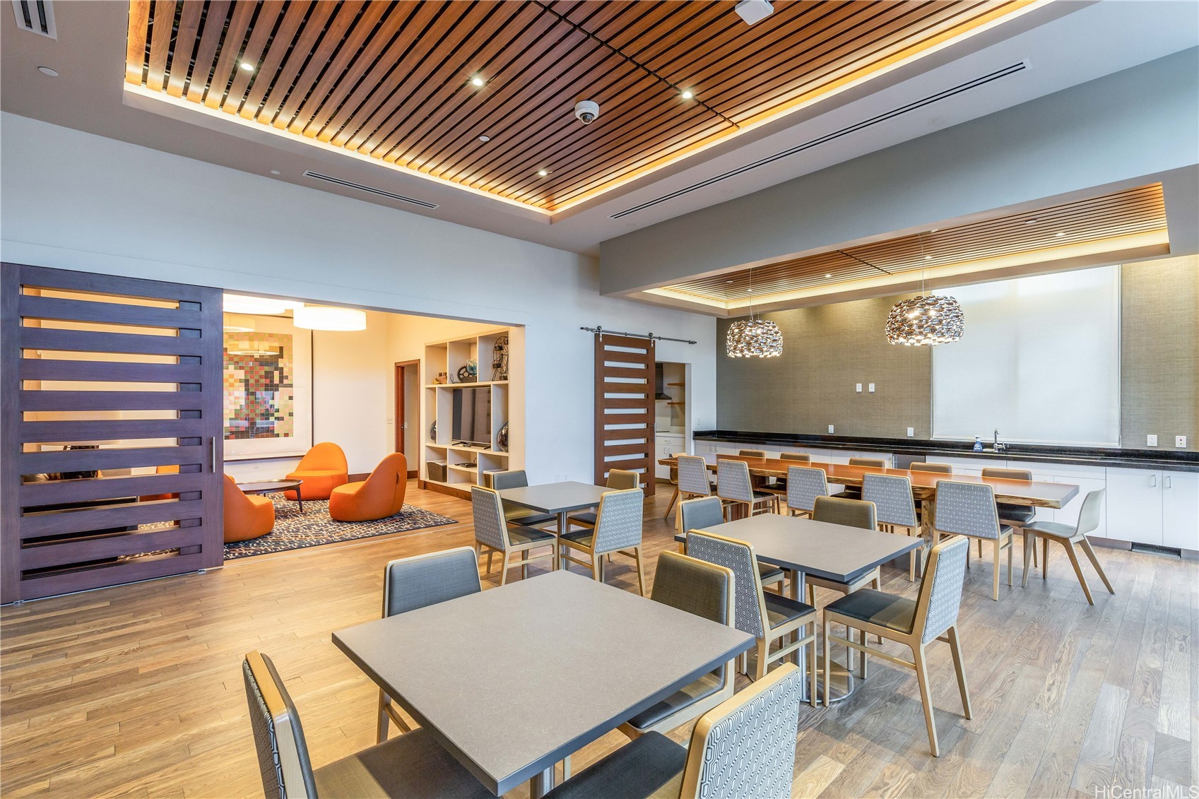 Furnished dining and lounge area with a slatted wood ceiling, kitchen counter, and orange lounge chairs.