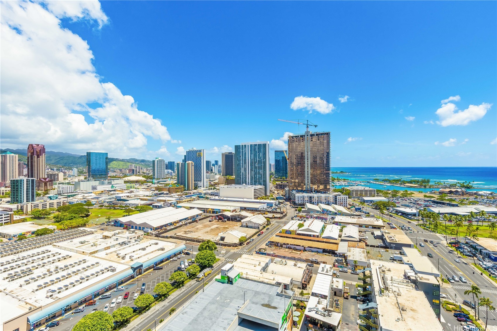 Aerial view of Honolulu’s high-rise district, harbor, coastline, and mountains under a bright sky.