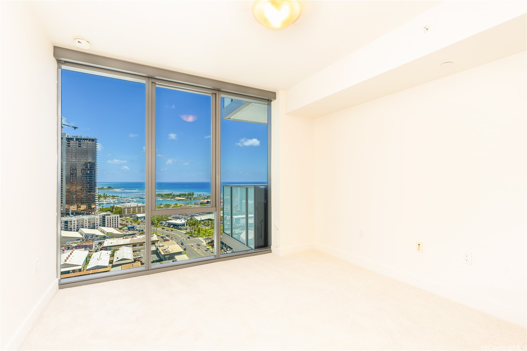 Empty carpeted condo room with floor-to-ceiling windows overlooking the ocean, harbor, and city.