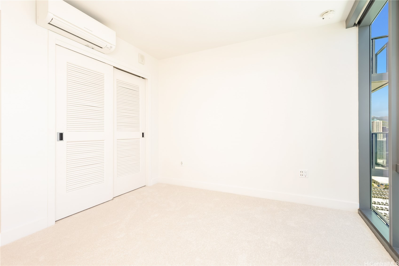 Empty carpeted room with louvered closet doors, wall-mounted air conditioner, and floor-to-ceiling city-view windows.