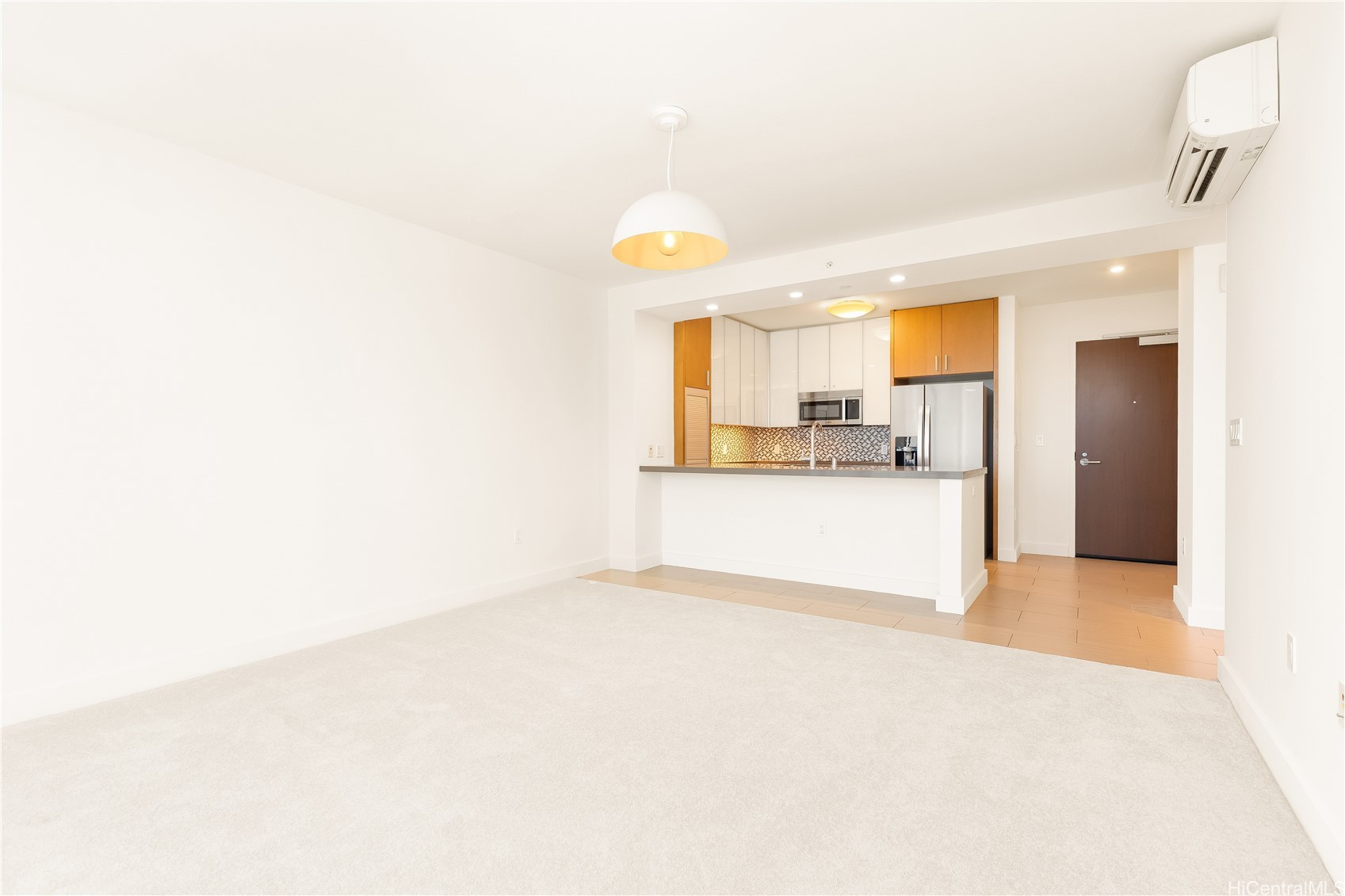 Vacant condo living room with carpet and an open kitchen featuring a breakfast bar and stainless steel appliances.