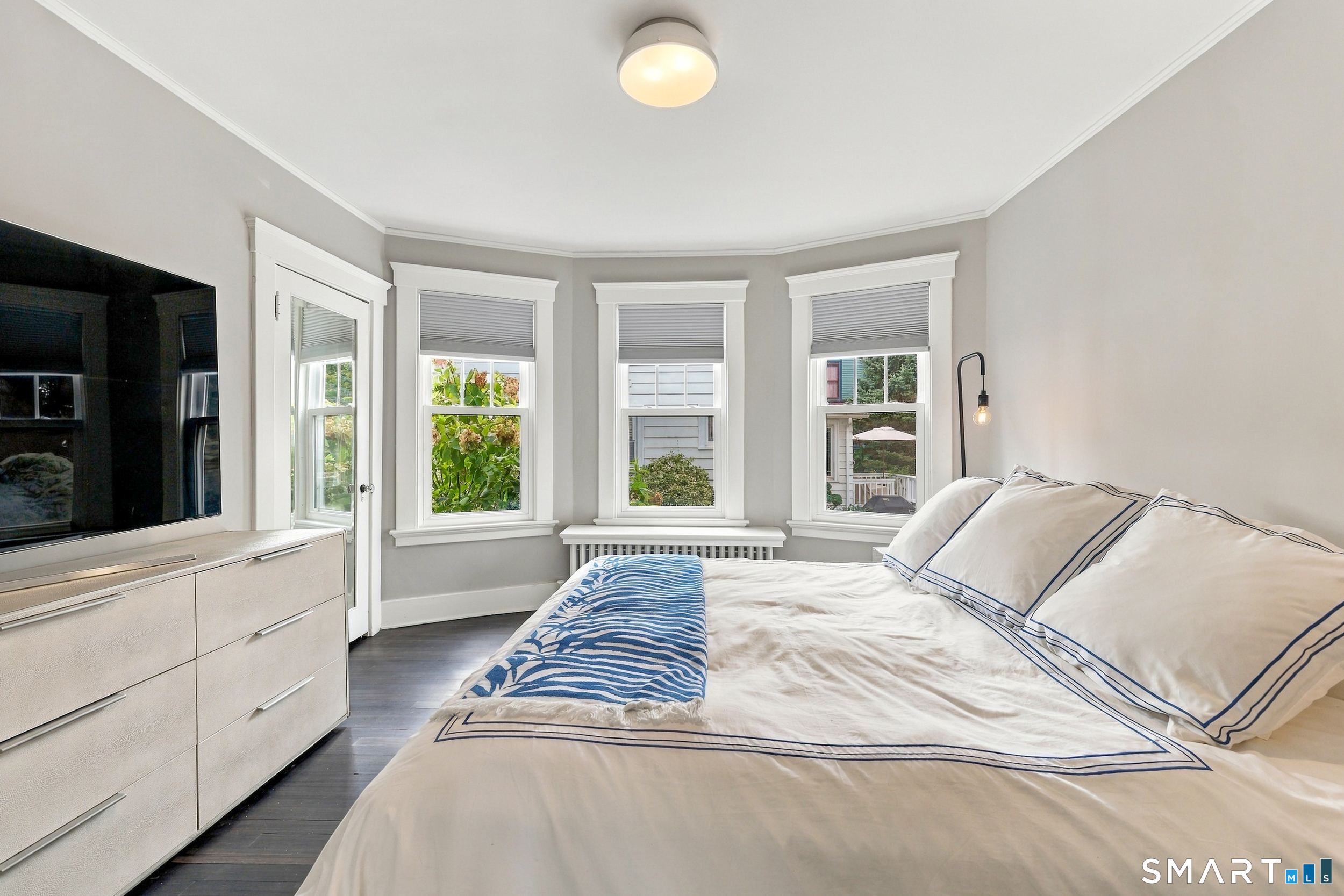 Bedroom with white-and-blue bedding, three windows, and a dresser with television.