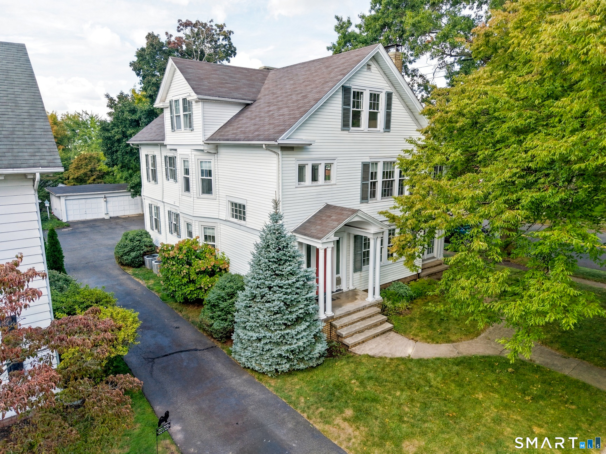 White-sided house with a columned front entrance, mature trees, driveway, and detached garage.