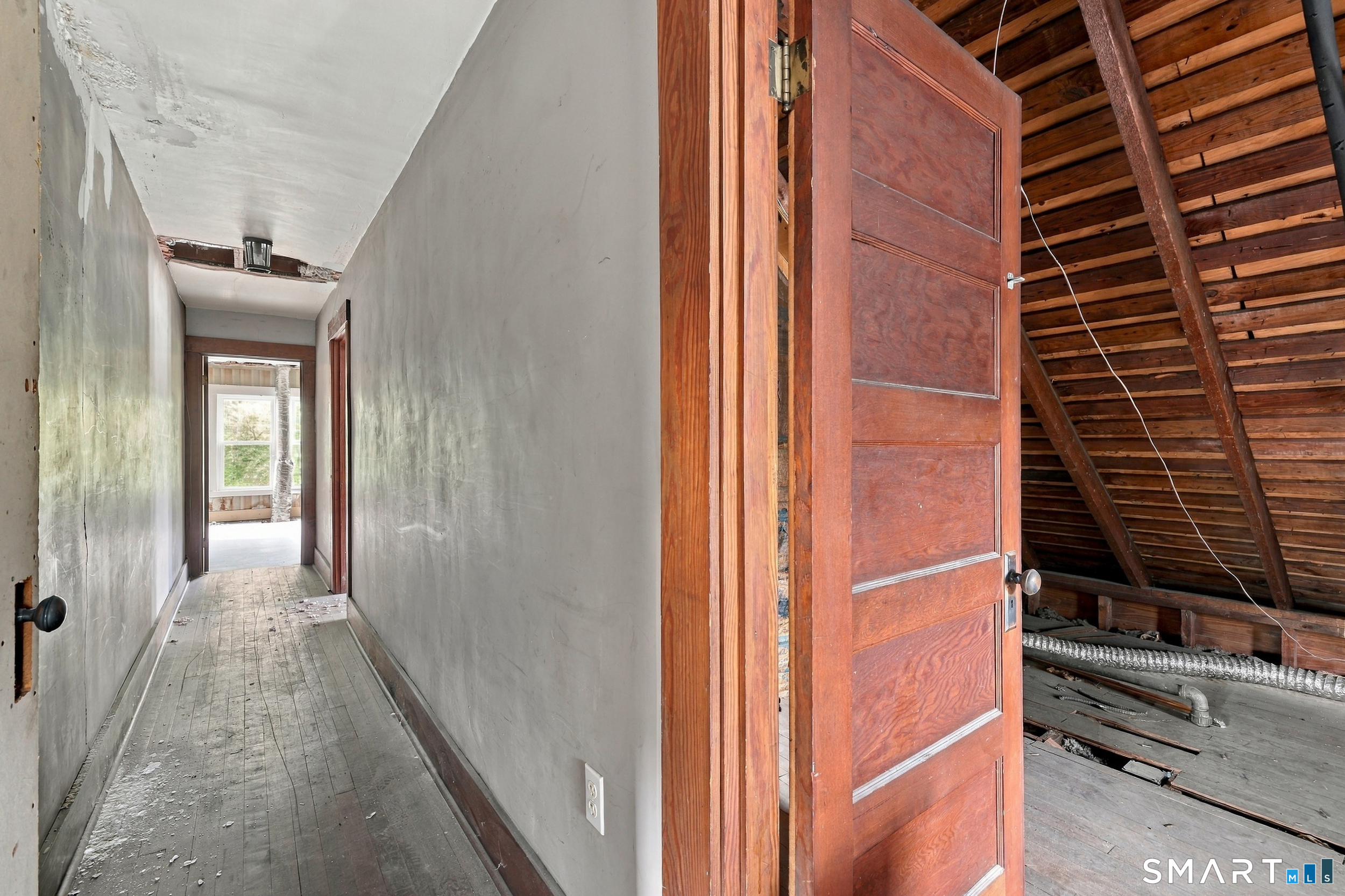 Unfinished hallway with worn wood floors and an open door to an attic with exposed rafters.