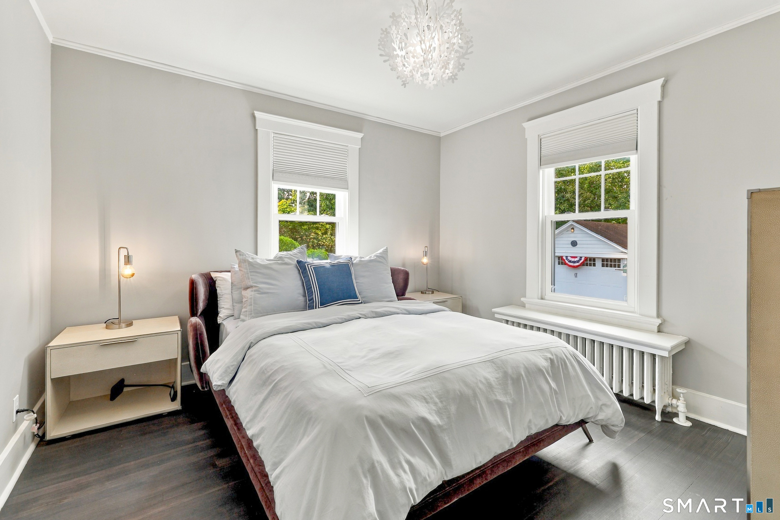 Staged bedroom with a white bed, blue accent pillow, and two tall windows above a radiator.