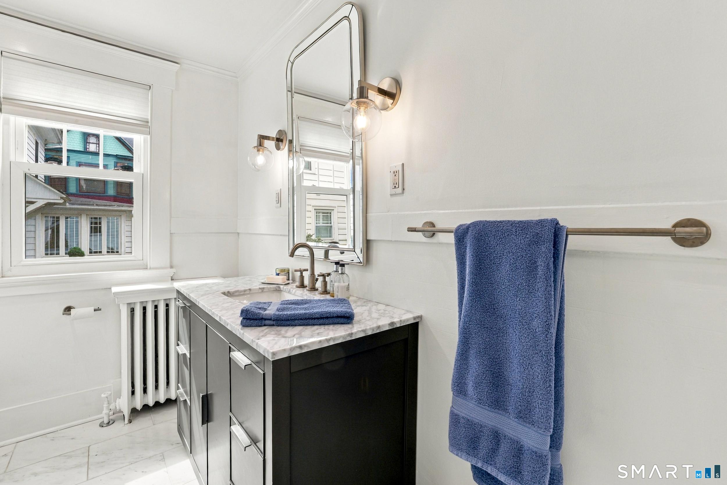 Bathroom with a dark vanity, marble surfaces, globe sconces, and blue towels beside a window.