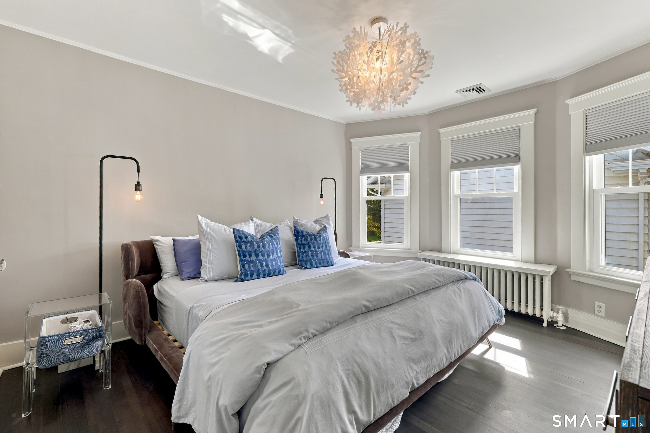 Staged bedroom with a large white bed, blue accent pillows, three windows, and a decorative chandelier.