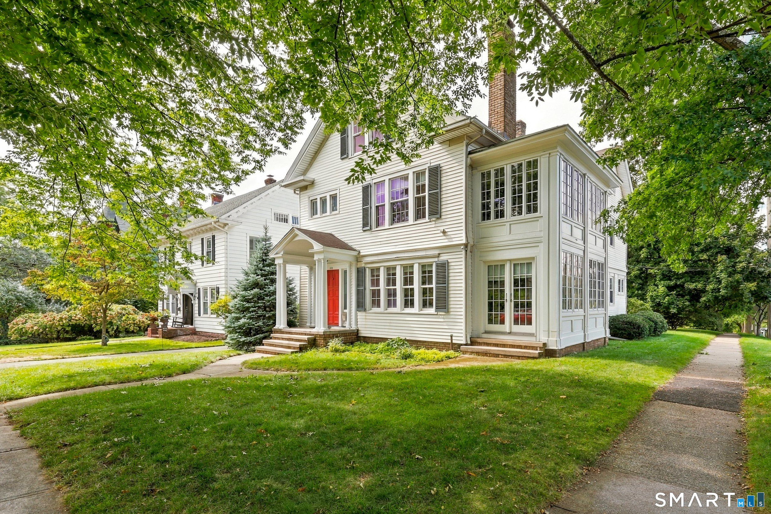 White multi-story house with a red front door, columned entry, and glass-sided sunroom beneath mature trees.