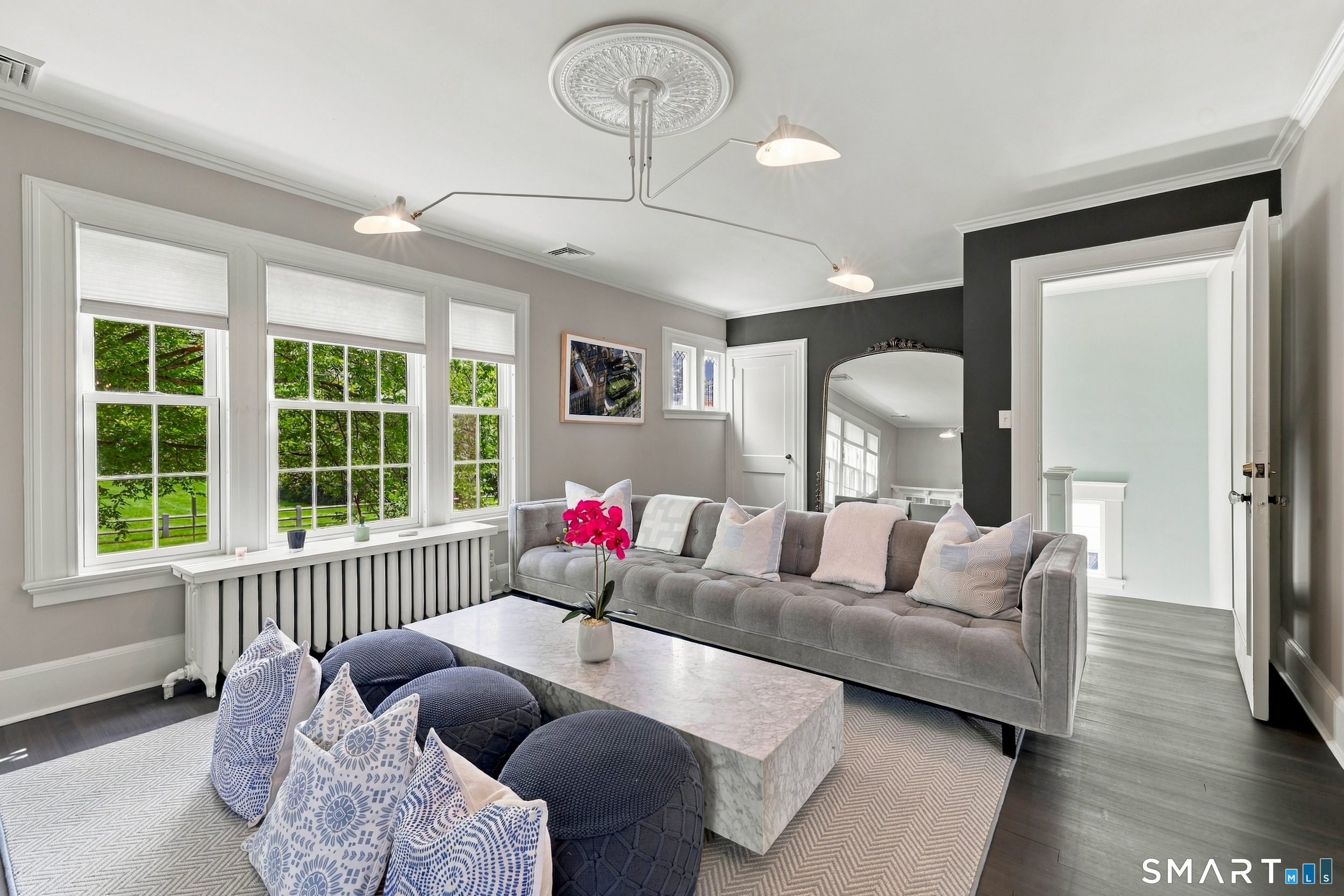 Living room with a gray sofa, marble coffee table, and three large windows overlooking greenery.