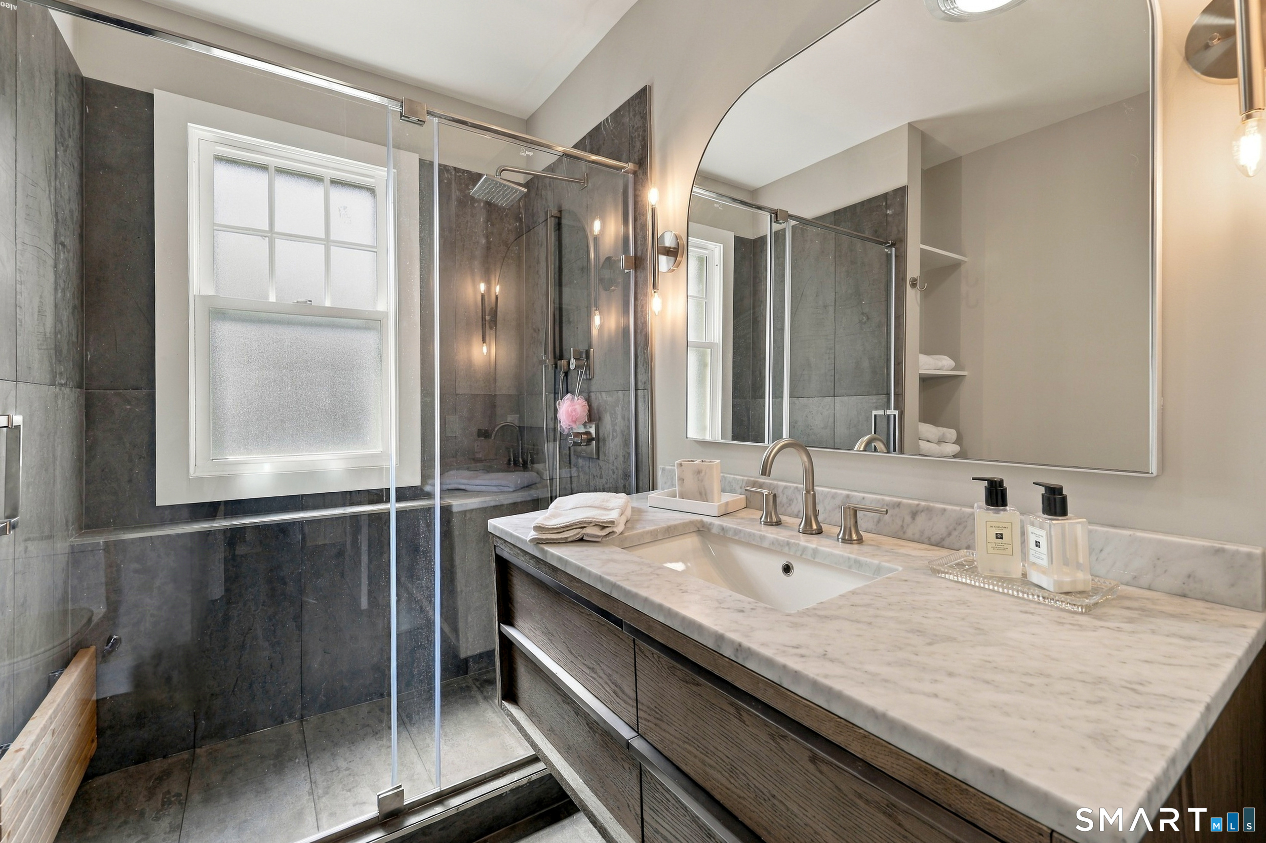 Bathroom with a glass shower, dark tile, frosted window, and marble-topped wood vanity.