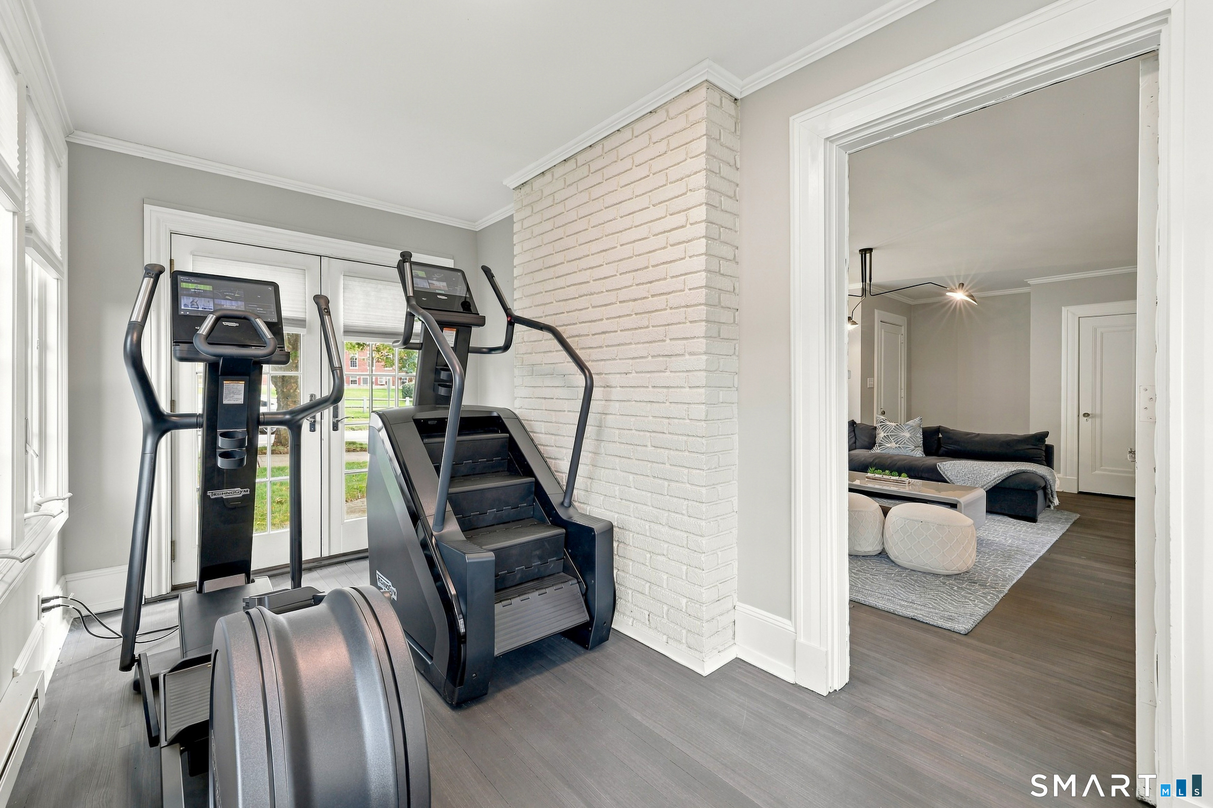 Home gym with cardio machines beside windows, opening onto a living room.