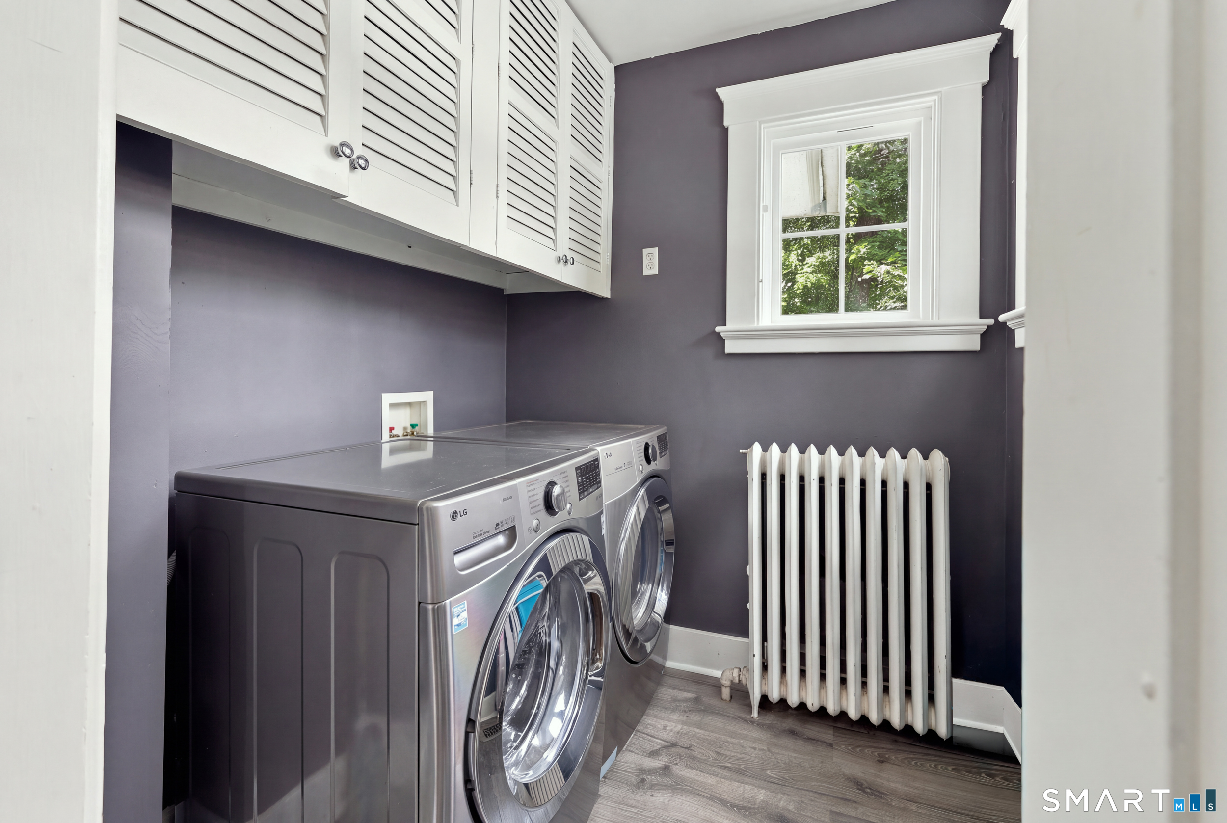 Laundry room with stainless-steel washer and dryer, white cabinets, and a radiator beneath a window.
